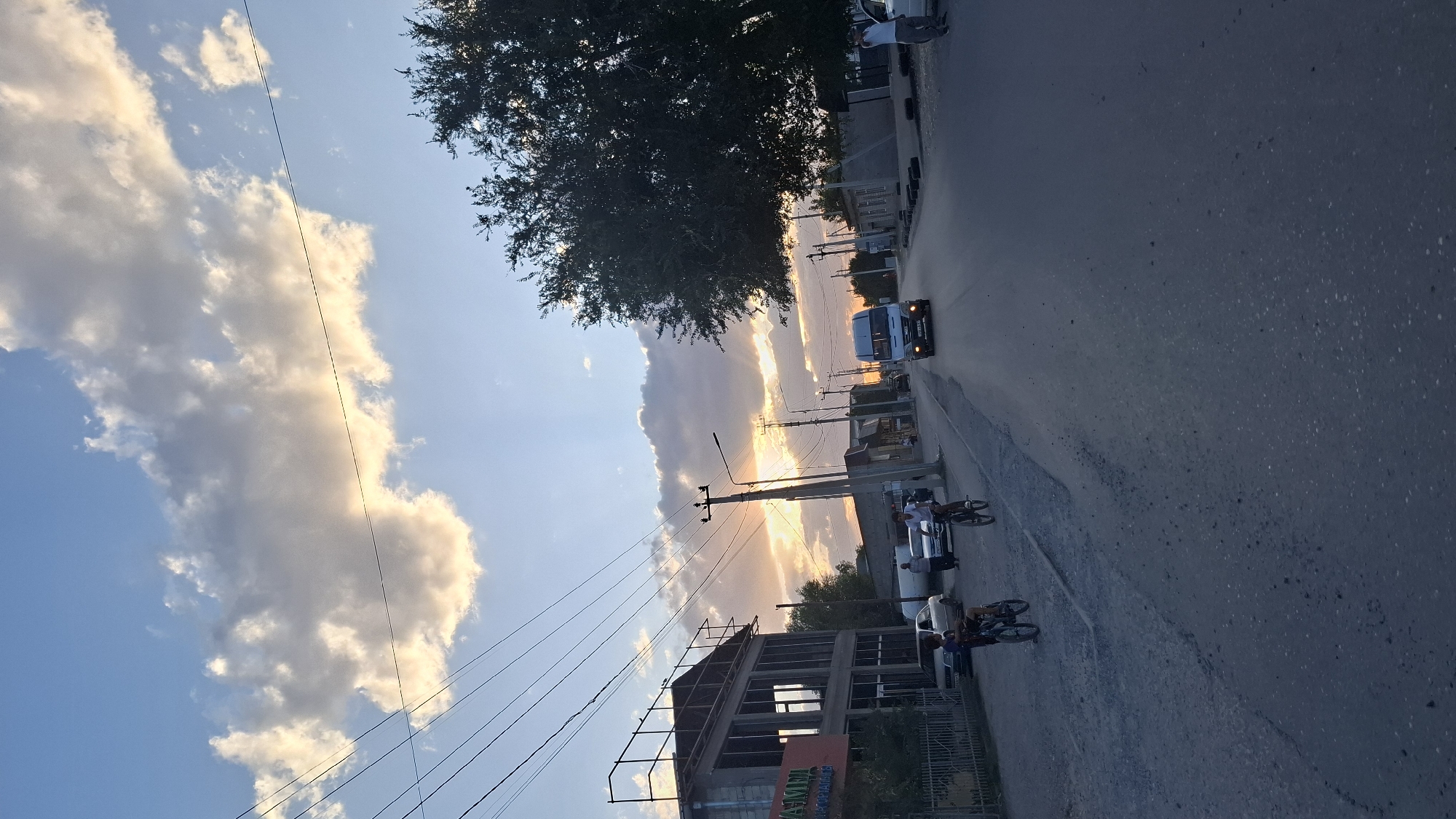 The image depicts a street scene in the city of Shubarssu, Kazakhstan, during sunset. The sky is filled with dramatic clouds, illuminated by the setting sun, which casts rays of light across the scene.
On the left side of the street, a partially constructed building with visible framing and windows stands. A fence encloses the property. Further down the street, several parked cars are visible. Two young individuals are riding bicycles on the side of the road.
The road itself is paved with asphalt, showing some signs of wear and tear. Power lines run overhead, supported by utility poles. A large tree with dense foliage is situated on the right side of the street.
In the distance, a white van is driving down the road. Other buildings and structures line the street, suggesting a mixed-use or residential area. A few pedestrians are also present, including one standing near the large tree on the right. The overall impression is of a typical evening in a town or city.