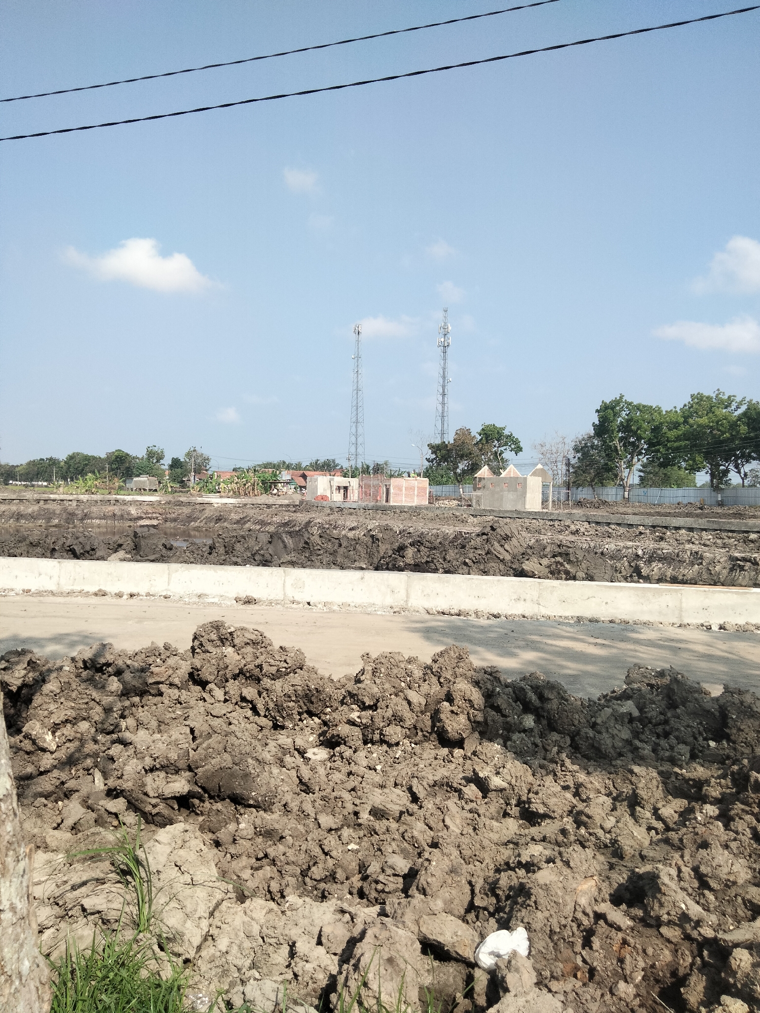 The image depicts an outdoor construction site in Sikampuh, Indonesia. In the foreground, a large pile of dark, clumpy soil is visible, with some green grass growing at the bottom left. A light-colored concrete barrier runs horizontally across the middle of the image, separating the foreground from the rest of the scene. Behind the barrier, a deep trench or excavation is filled with dark, wet mud and water.
Further in the background, there are several structures under construction, including brick walls and what appear to be foundations for buildings. Two tall cell towers are prominent against the clear blue sky. Trees and scattered buildings are visible on the horizon, suggesting a rural or semi-urban environment. Two black wires are strung across the top of the image. There are no people visible in the scene. The overall impression is one of ongoing development and earthworks.