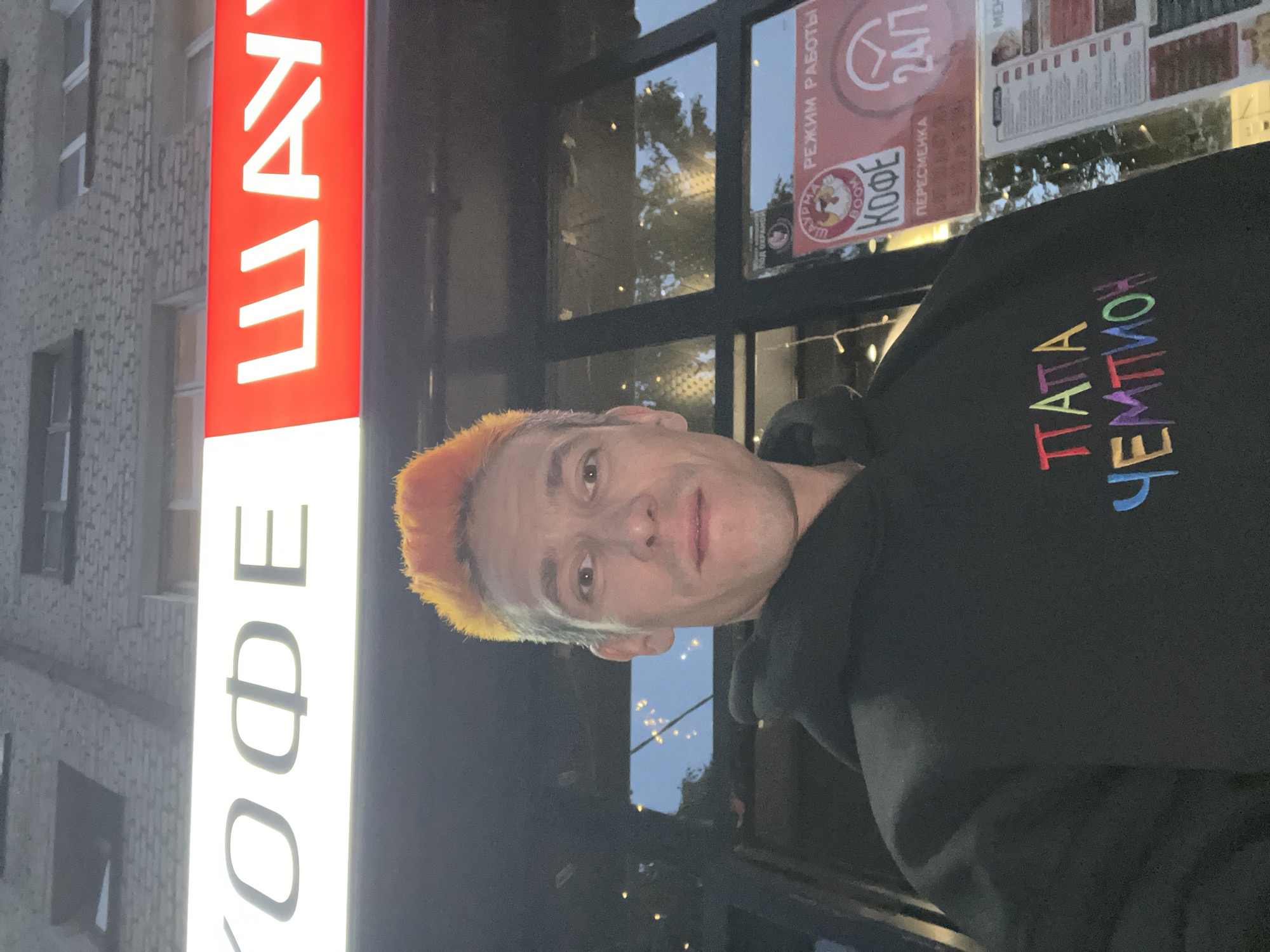 An individual is depicted in a medium shot, centered in front of a commercial establishment in the city of Chaikovsky, Russia. The person, an adult male, has short, spiky hair, predominantly bright orange on the top with lighter sides, possibly graying. He wears a black hooded sweatshirt with multicolored text on the chest that reads "ПАТТА ЧЕМПИОН" (PATTA CHEMPION). His gaze is directed towards the camera with a neutral expression.
Behind the individual, a brightly illuminated storefront is visible. Above the main entrance, a prominent sign displays services: the left section, with a white background and dark lettering, partially reads "КОФЕ" (KOFE, meaning Coffee). The right section, with a red background and white lettering, partially reads "ШАУ" (SHAU, likely from SHAURMA, meaning Shawarma).
Below the main sign, through a large glass window, additional signage and interior reflections are discernible. A circular logo featuring a cartoon chicken with the text "ШАУРМА BOOM" (SHAURMA BOOM) and "КОФЕ" (KOFE) is present. Adjacent to this, a red rectangular sign displays "РЕЖИМ РАБОТЫ" (REZHIM RABOTY, meaning Operating Hours) with a "24/7" indicator and a clock icon, signifying round-the-clock service. The word "ПЕРЕСМЕНКА" (PERESMENKA, meaning Shift Change) is also visible. To the left, a small icon with Cyrillic text that appears to be "ПОД ОХРАНОЙ" (POD OKHRANOY, meaning Under Protection/Security) can be seen. A menu board with various items is partially visible to the right. The upper background shows a brick building facade with windows. The scene is illuminated by artificial light, suggesting evening or nighttime.
