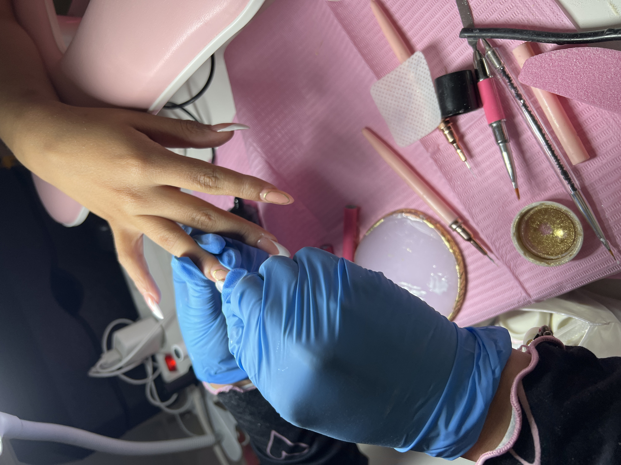 The image depicts a close-up, overhead view of a nail technician, wearing blue nitrile gloves, working on a client's fingernails. The client's hand is resting on a pink manicure table, with the technician meticulously applying product to one of the nails. Various nail art supplies, including brushes, a small pot of glitter, a nail file, and a palette, are scattered across the pink table mat. A pink nail lamp is partially visible in the background. The scene appears to be set in a nail salon. There is no indication of the specific location within Madrid, Spain.