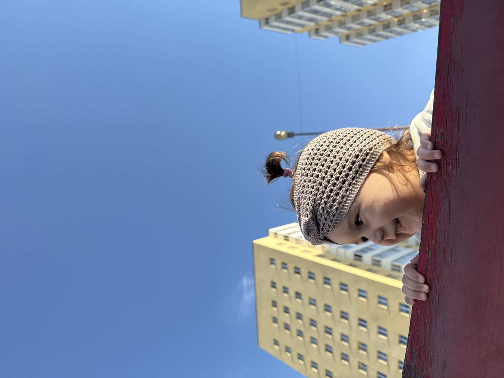 A low-angle, medium shot shows a young child peeking over a red wooden structure against a bright blue sky. The child, wearing a beige knitted cap with a small ponytail, is smiling and gripping the edge of the structure with their hands. In the background, two tall buildings are visible, one with a light yellow facade and the other with a white facade. A street lamp can be seen between the two buildings. The location is Tobolsk, Russia.