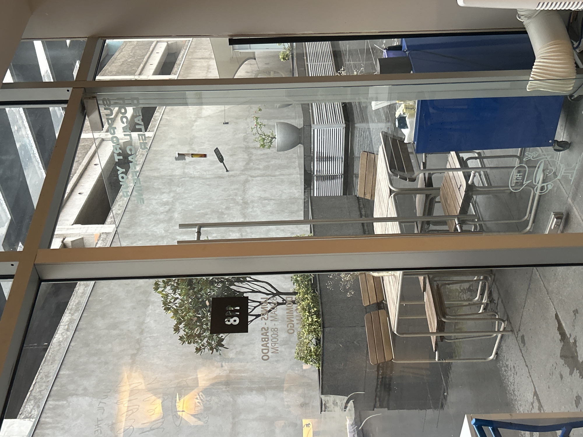 The image depicts a view through a glass window, possibly of a restaurant or cafe, in San Bernardino Tlaxcalancingo, Mexico. Outside the window, there are tables and chairs arranged on a patio or sidewalk. The furniture appears to be made of metal and wood. There is a potted plant and some greenery visible as well. Reflected in the glass are some written texts and a sign with the number "8". The outside environment includes an adjacent building, and a glimpse of the street.