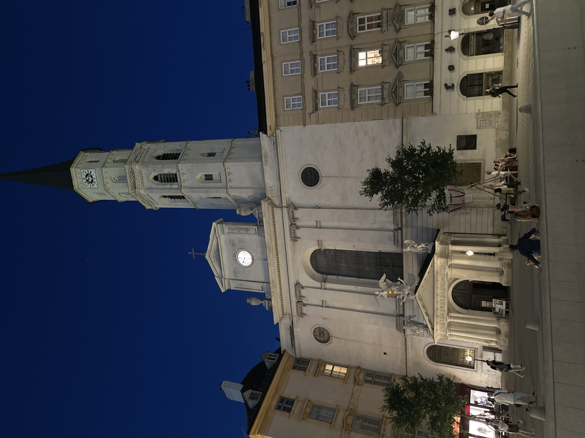 This media file depicts a nighttime scene in Vienna, Austria, showcasing a prominent church with a tall, ornate tower and a larger, lighter-colored building adjacent to it. The sky is a deep, clear blue, indicating a dry evening.
The church tower, made of white stone, features a clock face with Roman numerals and is topped with a dark, pointed spire. Decorative architectural elements, including arched windows and intricate carvings, are visible on the tower. The building next to the church is also made of light-colored stone, with multiple floors and numerous windows, some of which are illuminated.
Several people are present in the scene. On the sidewalk in front of the lighter building, a group of individuals are gathered, appearing to be seated or standing. Further along the street, more people are walking or standing, some interacting with each other. One person is seen leaning against a railing, looking out.
The activity appears to be a casual evening gathering or stroll. The lighting suggests it is after sunset, with artificial lights illuminating the buildings and street. The overall mood seems calm and relaxed. No visible text is present in the image.