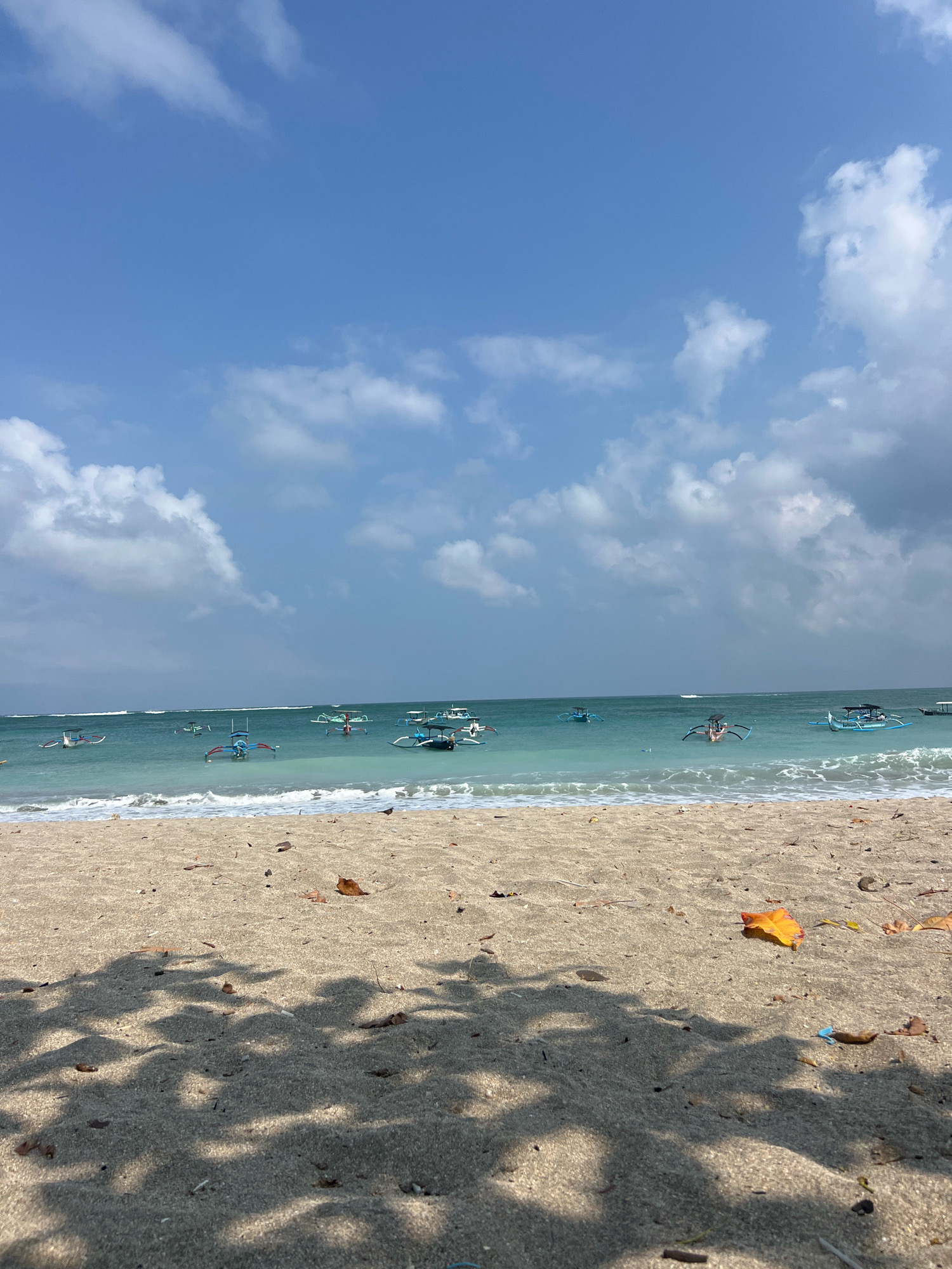 A low-angle, eye-level shot captures a serene beach scene in Kuta, Indonesia. The foreground is dominated by a sandy beach, dappled with the shadows of leaves from an unseen tree overhead. A few scattered dry leaves, including a vibrant orange one, lie on the sand. In the midground, gentle waves roll onto the shore, creating a white foam line. The turquoise water of the ocean stretches to the horizon, dotted with numerous traditional outrigger boats, some with colorful canopies, bobbing gently on the surface. In the background, a vast expanse of blue sky is filled with scattered white cumulus clouds, some appearing wispy and others more defined. The overall impression is one of a peaceful, sunny day at a tropical beach.