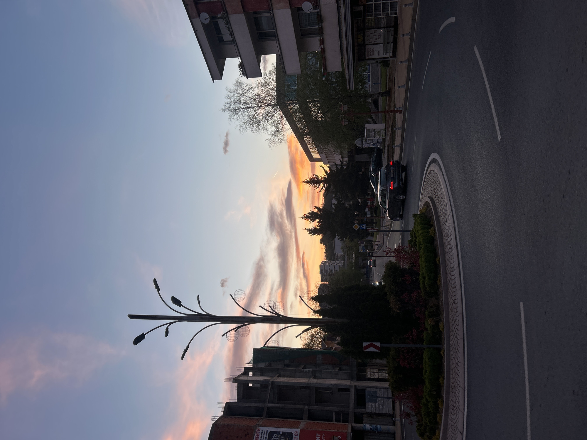 The image depicts a street scene in Zlatograd, Bulgaria at twilight. The sky features streaks of orange and pink clouds against a blue backdrop. An unfinished building of brick and concrete is in the lower foreground. A decorative lamppost with spherical ornaments stands near the building. Beyond the lamppost are trees and buildings, and a road with a roundabout. Two dark-colored vehicles are parked near the curb. The building and street indicate an urban setting.
