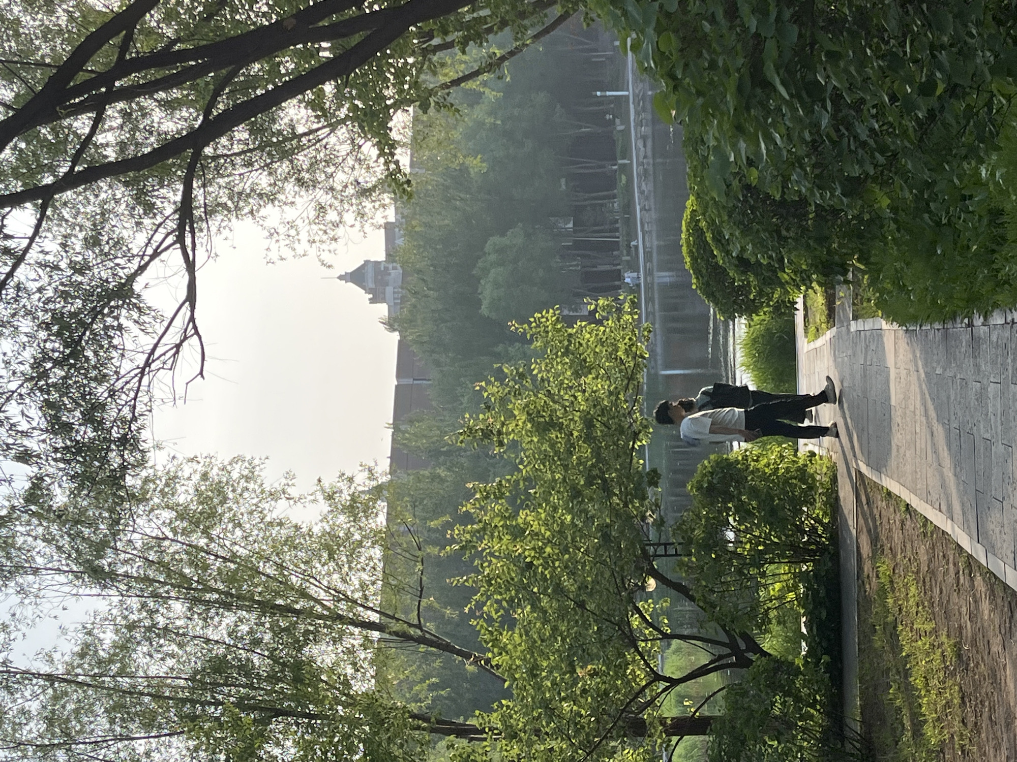 The image shows a park scene in Changchun City, China. A brick walkway curves along the edge of a body of water, possibly a lake or river, which reflects the surrounding trees and sky. Two figures, possibly men, walk along the path. Dense foliage, including trees and bushes, lines both sides of the walkway. A building with a pointed roof is visible in the background, shrouded in the misty atmosphere. The sky is bright and hazy.