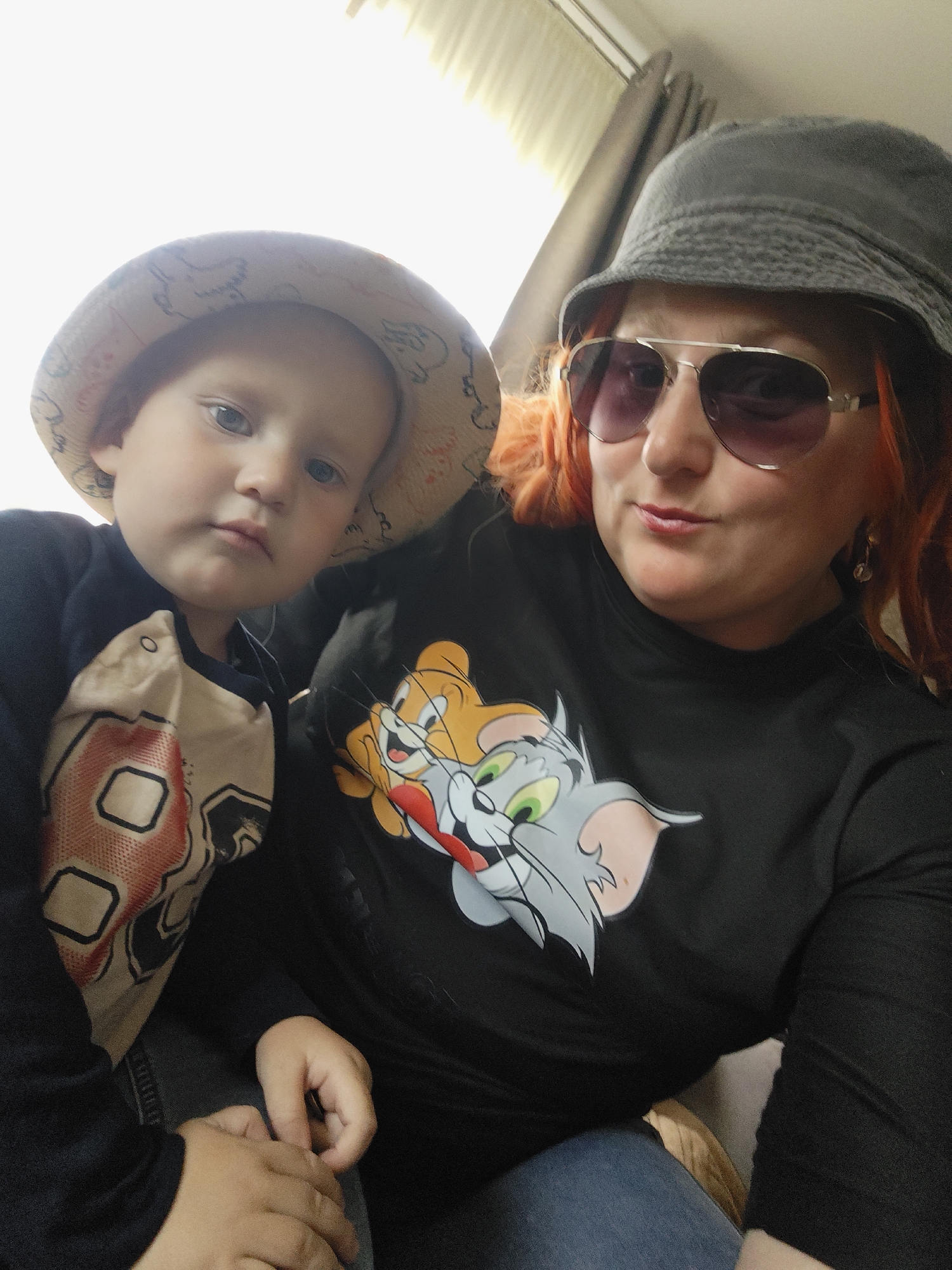 The image shows a selfie taken indoors, likely in an apartment in Mokino, Russia. A woman with bright orange hair, wearing a gray bucket hat and aviator sunglasses, is posing next to a young child who is wearing a patterned bucket hat. Both are looking at the camera. The woman is wearing a black t-shirt with a graphic print of Tom and Jerry characters. The child is wearing a dark long-sleeved shirt with a tan shirt layered underneath. The background shows a window with blinds and a curtain. The lighting is natural and bright, suggesting daytime.