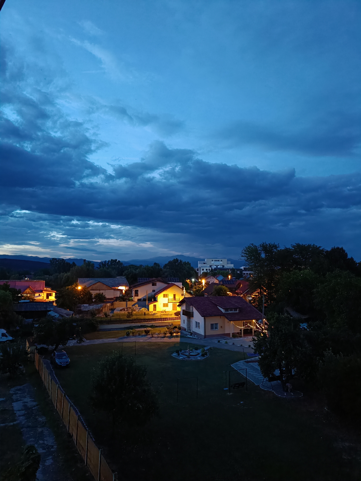 The image captures an aerial view of a residential area in Târgu Jiu, Romania, at dusk. The sky is a deep blue, filled with dramatic, dark clouds that suggest an approaching storm. In the foreground, a well-maintained yard is visible, featuring a swing set and a paved path. A wooden fence runs along the left side of the property, separating it from a concrete area. A dark-colored car is parked in the driveway.
Several houses are illuminated with warm, artificial lights, creating a cozy contrast with the darkening sky. One house in the center of the frame has a bright yellow glow emanating from its windows and porch lights, with decorative string lights draped across its facade. Another house to the left, with a red roof, also has illuminated windows. In the background, more houses and trees are silhouetted against the twilight sky, with a larger, modern building visible further in the distance. The landscape beyond the residential area shows a range of rolling hills and mountains, their forms softened by the dim light. The overall atmosphere is tranquil yet tinged with the anticipation of evening or possible bad weather.