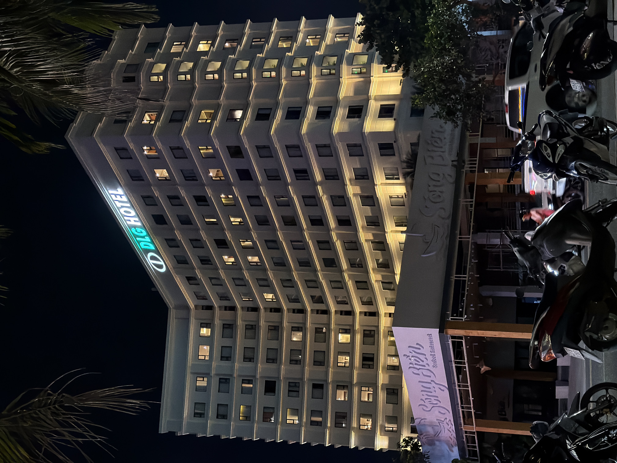 This is a night exterior shot of the DLG Hotel in Da Nang, Vietnam. The hotel is a tall, white building with many lit windows, indicating occupancy. Neon green signage on the top of the building reads "DLG HOTEL". Below the hotel, the "Song Bien Seafood Restaurant" is visible, with its name displayed in large white lettering on a signage board. Several motorcycles are parked in the foreground, with a white pickup truck and another vehicle visible in the background. Palm fronds hang from the top left of the image, suggesting a tropical location. The overall scene captures the lively atmosphere of a city at night.