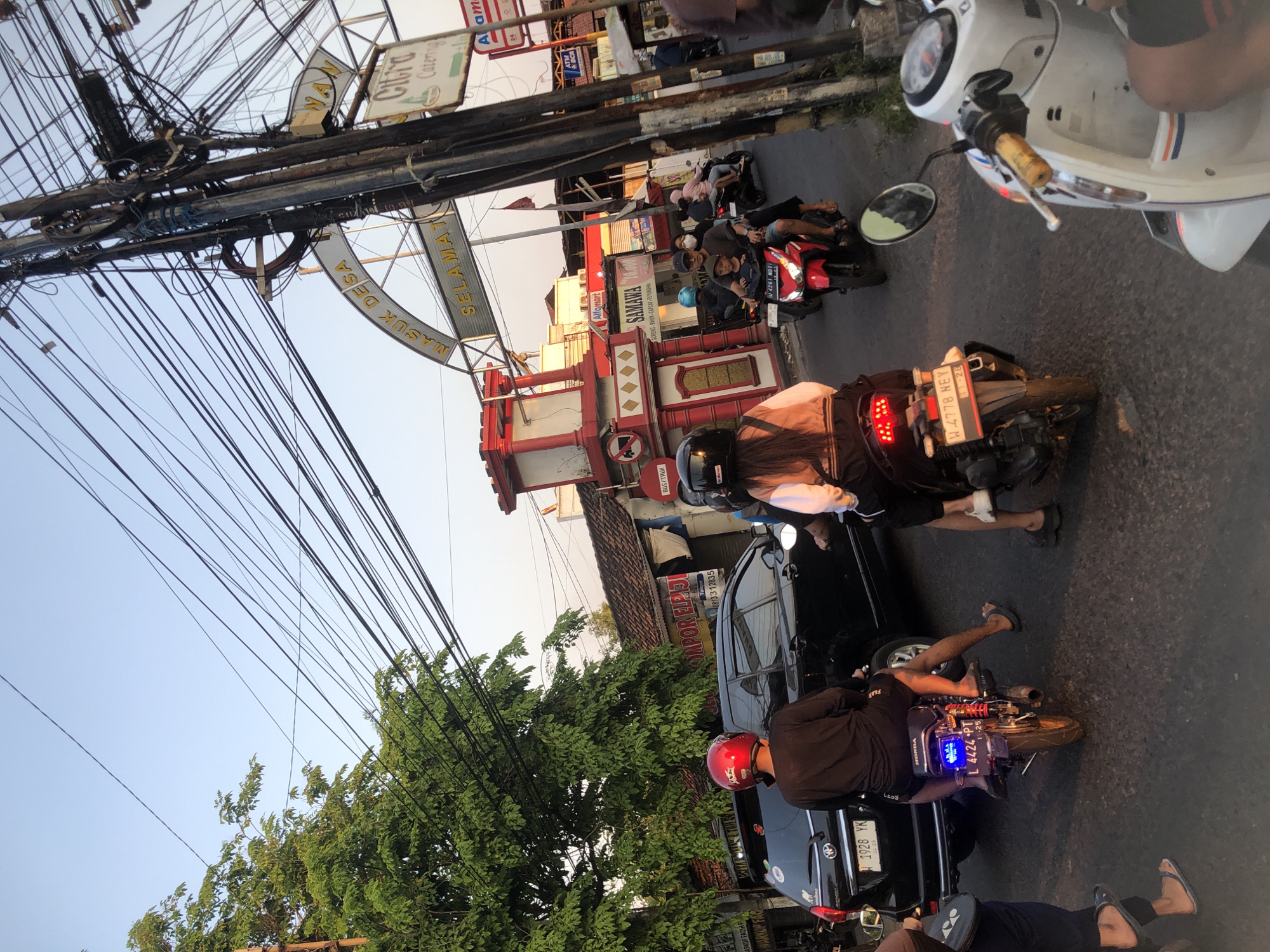 The image captures a street scene in Tenggulunan, Indonesia, characterized by a busy intersection with several motorcycles and a car. The time of day appears to be late afternoon, with the warm glow of sunset visible in the sky. Overhead, a dense network of electrical wires crisscrosses the frame, a common sight in Indonesian urban landscapes.
In the foreground, a person on a motorcycle wearing a red helmet is about to enter the intersection. To their right, a black car is stopped, and behind it, another motorcycle with two passengers navigates through the traffic. Further down the road, more motorcycles are visible, some with passengers, suggesting a bustling commuter environment.
A distinctive red and white archway, adorned with the words "MASUK DESA SELAMAT DATANG," marks the entrance to a village, providing a geographical marker for the location. Various storefronts and signs, including "Alfamoart" and "Citro Catering," indicate commercial activity in the area. The road surface is asphalt, showing signs of wear and tear, common in local infrastructure. The overall impression is one of a lively, everyday scene in a developing urban area.