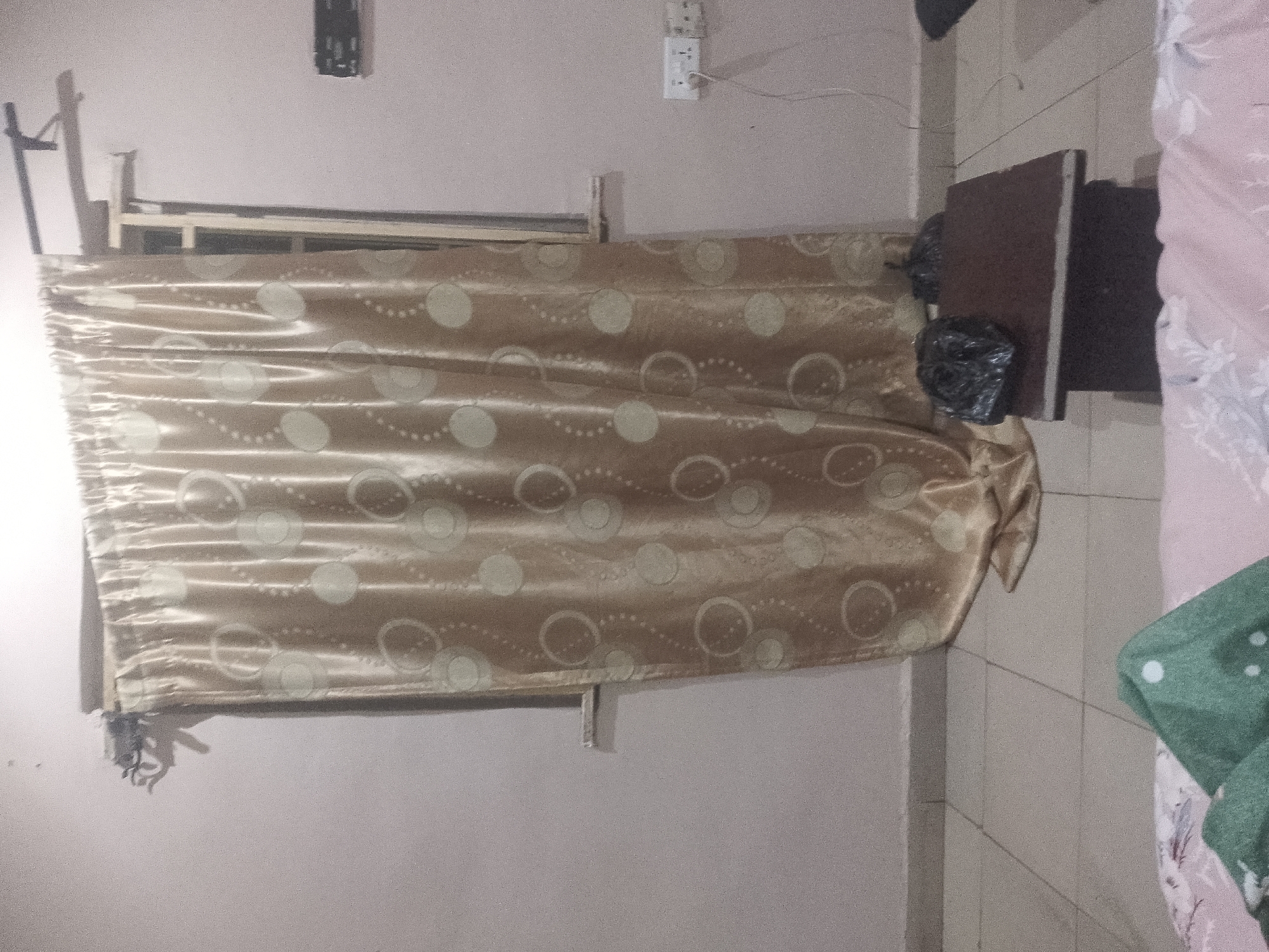 A head-on, medium shot captures a beige curtain with circular patterns hanging across a wall. To the left, a thin metal rod extends vertically from a horizontal bar above the curtain. To the right of the curtain, a dark brown, rectangular object, possibly a small table or shelf, stands against the wall. Two black plastic bags are placed on the surface of this object. A pink, patterned fabric is visible to the far right, partially obscuring a green fabric item. The wall has a textured finish.