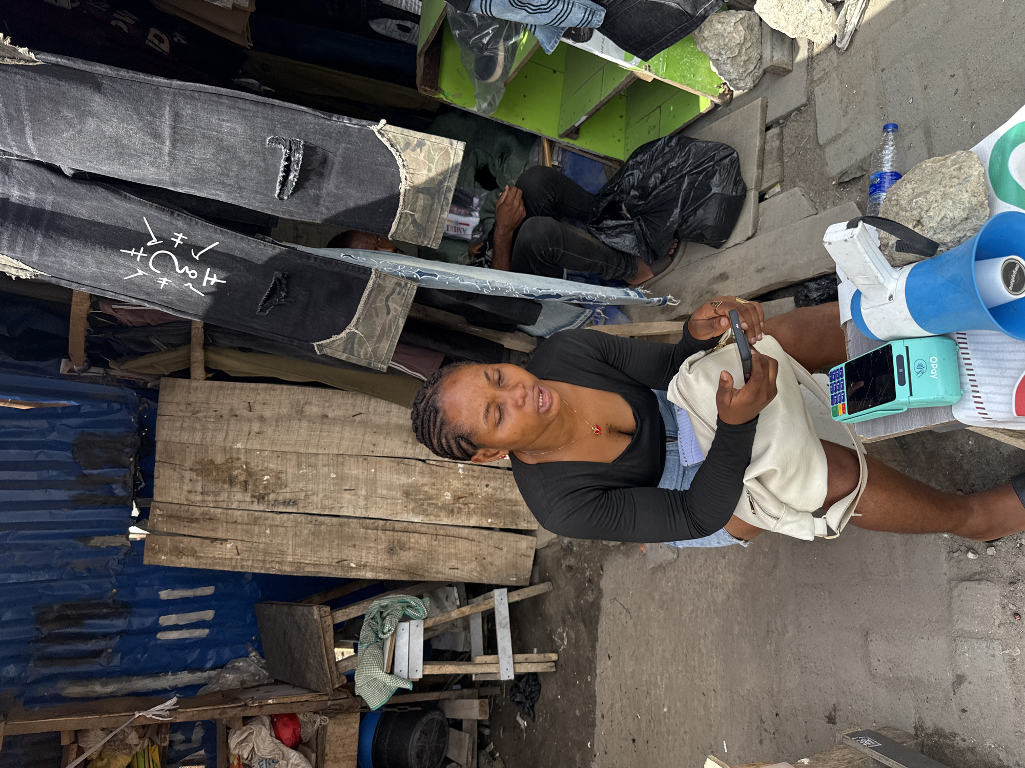 The image depicts a scene in Lagos, Nigeria, with a woman sitting near a makeshift stall or shop. The stall has a blue corrugated metal wall and is constructed from wooden planks. The woman, wearing a black long-sleeved shirt and denim shorts, sits facing the camera. She has braided hair and is holding a smartphone in her hands. Next to her is a POS machine, a megaphone, a bottle of water, and a stone. A pile of jeans, some with stylized patterns, are displayed on the left. Behind the jeans, a man is lying down. The environment appears cluttered, suggesting a busy market or street-side location.