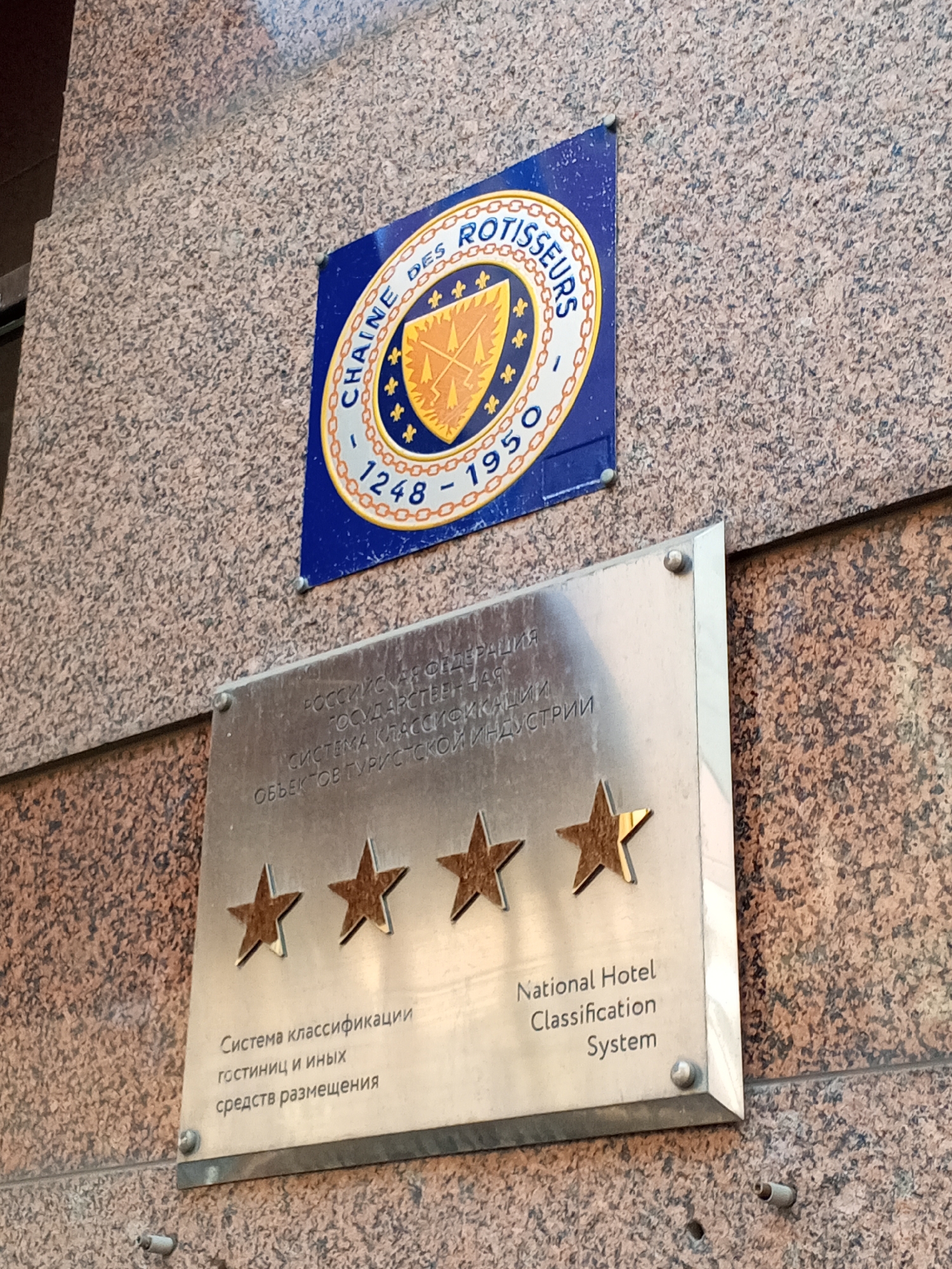 The image displays two distinct plaques mounted on a mottled, light-colored stone wall, located on a building in Saint Petersburg, Russia. The upper plaque is a weathered blue square featuring a circular golden emblem with three crossed arrows, encircled by the text "CHAÎNE DES ROTISSEURS" and the dates "1248 - 1950." Below it, a rectangular, reflective metallic plaque is affixed to the wall with four visible silver fasteners. This lower plaque contains text in both Cyrillic and English, identifying it as a "National Hotel Classification System." It prominently displays four gold-colored stars arranged horizontally, suggesting a four-star rating, with the position for a fifth star either empty or obscured at the far right. Below the lower plaque, a small, exposed metal anchor protrudes from the wall, adjacent to an empty drill hole. No people, specific street addresses, or active interactions are visible.
