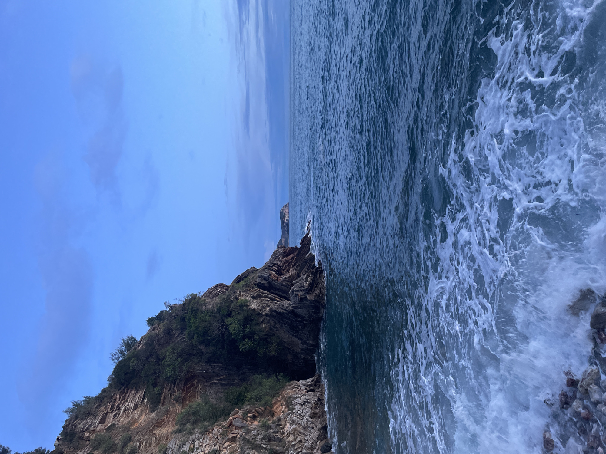 The scene depicts a natural coastal landscape in Rafailovići, Montenegro, under ambient dim light, consistent with twilight or dawn. A substantial rocky cliff, characterized by visible geological strata and sparse green vegetation along its ledges and crest, dominates the left side of the frame, extending from the foreground into the upper left. The cliff face plunges directly into the sea. The sea occupies the majority of the lower and right portions of the image, presenting as dark blue with moderate surface agitation. White foam patterns are visible in the immediate foreground, indicative of waves breaking against unseen or partially visible rocks at the base of the cliff. The horizon line separates the agitated sea from a light blue sky with scattered, faint cloud formations. A distant landmass or island is discernible on the horizon toward the far left of the image. No human individuals, animals, or man-made structures are present in the immediate foreground or midground.