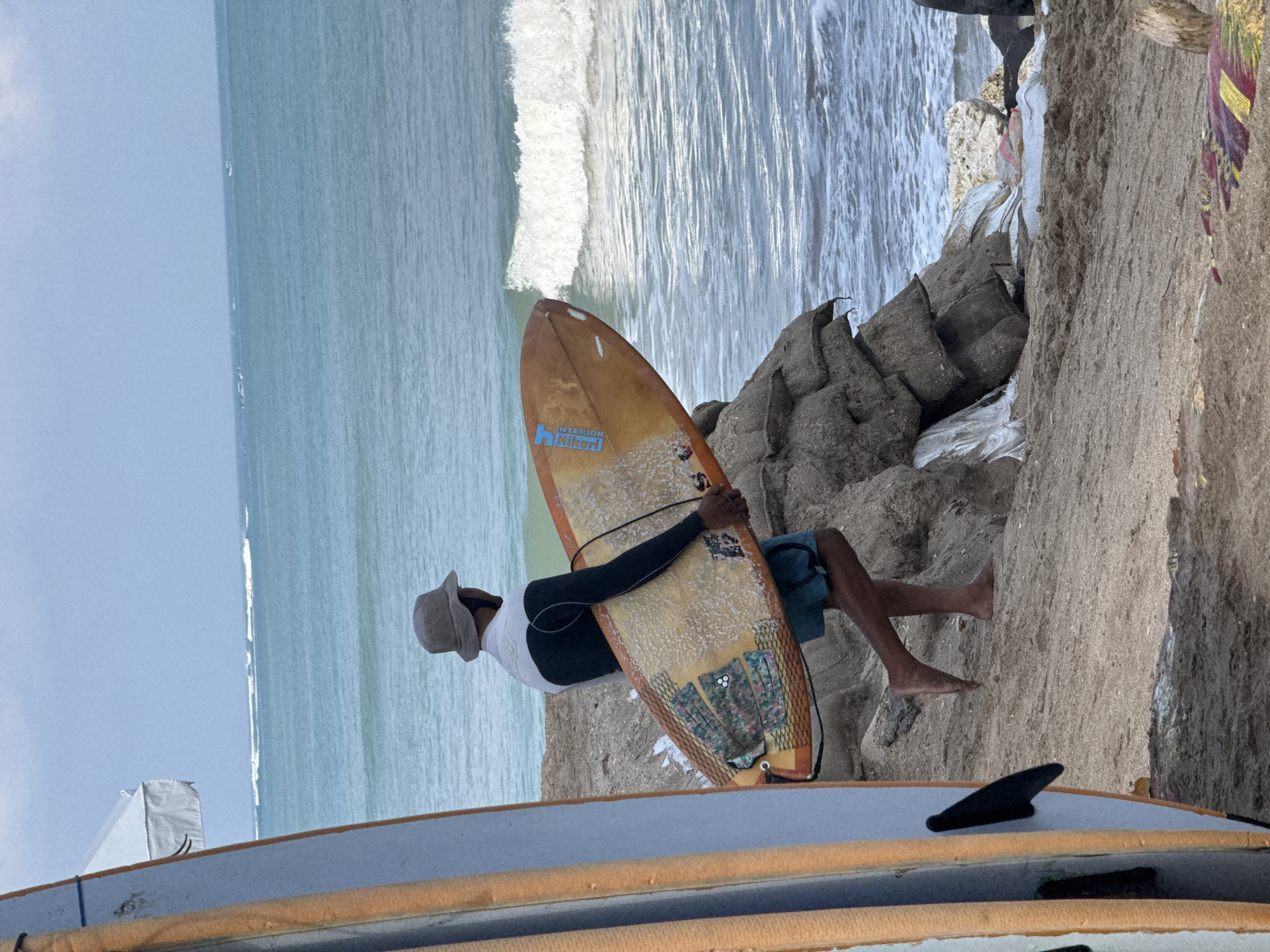 A full-shot, outdoor, eye-level, vertical photograph captures a surfer walking along the beach in Kuta, Indonesia. The surfer, positioned in the mid-right of the frame, is a person with dark skin wearing a gray bucket hat, a black and white long-sleeved rash guard, and blue swim trunks. They are carrying a light orange surfboard under their right arm. The surfboard has several traction pads on its surface. To the left of the surfer, another surfboard, predominantly light blue with orange accents and black fins, is visible, partially obscuring the surfer's left side.
The environment is a sandy beach with a rocky shoreline and a clear blue ocean in the background. Waves are breaking on the shore, and the sky is overcast with a pale blue hue. In the mid-ground, a pile of sandbags, likely used for coastal erosion control, is situated on the beach, creating a rough, textured barrier. The overall lighting suggests an afternoon or late morning scene. The photograph is taken from a medium distance, focusing on the surfer and their immediate surroundings.