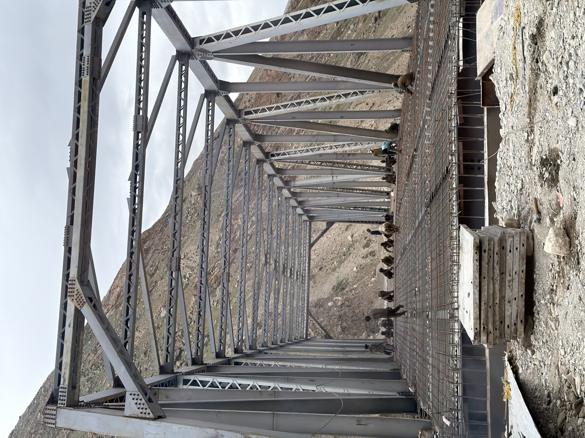The image shows the construction of a steel truss bridge over a rocky, mountainous terrain. The bridge structure is largely complete, with workers on the deck preparing for concrete pouring. The deck is covered with a grid of rebar, and several workers are visible sitting or standing on it. In the foreground, a stack of precast concrete slabs is piled on the ground. The background features steep, barren mountain slopes under a cloudy sky. The scene appears to be in a remote, possibly underdeveloped area, indicated by the rough terrain and the method of construction. There are approximately 15-20 individuals visible, appearing to be construction workers, engaged in various tasks related to the bridge construction. The overall environment is dusty and rocky, consistent with a construction site in a mountainous region.