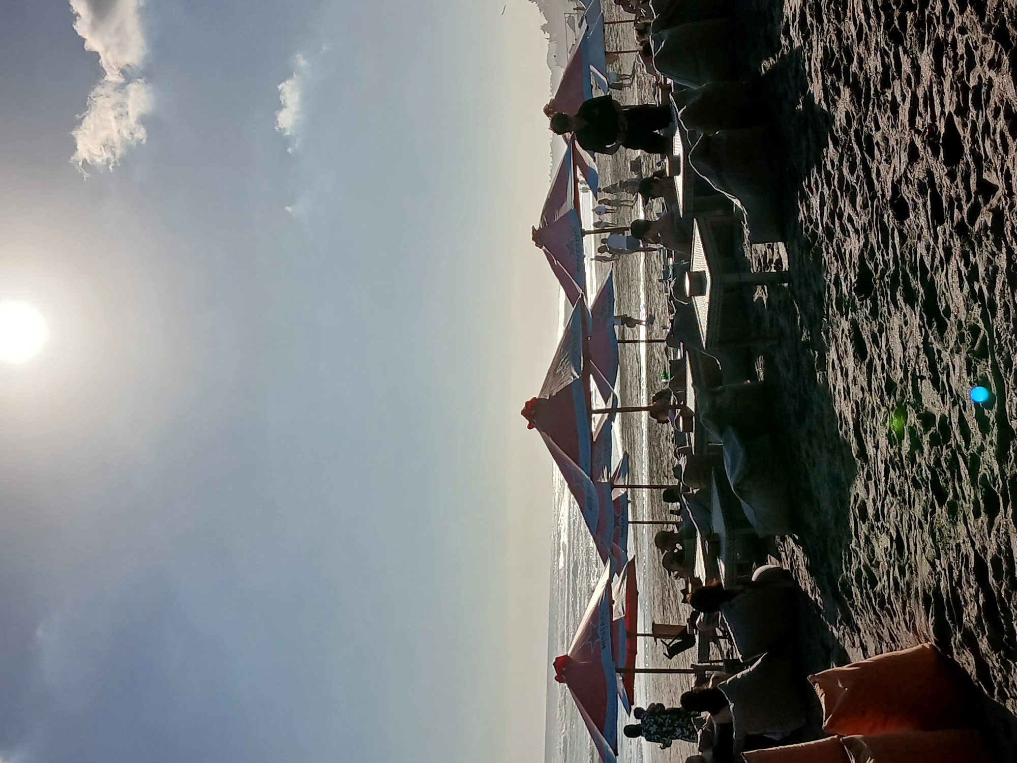 The image depicts a crowded beach scene in North Kuta, Indonesia, likely on a sunny day with moderate waves. In the foreground, sandy ground is covered with beanbag chairs and small tables, some with patrons seated. In the middle ground, several large, red and blue umbrellas provide shade over more tables and seating. A man in a dark shirt stands near a table. Further back, more people are visible walking along the beach and by the water's edge. The ocean stretches to the horizon, meeting a bright sky with a prominent sun and scattered clouds. The overall atmosphere suggests a relaxed and bustling beachside establishment.