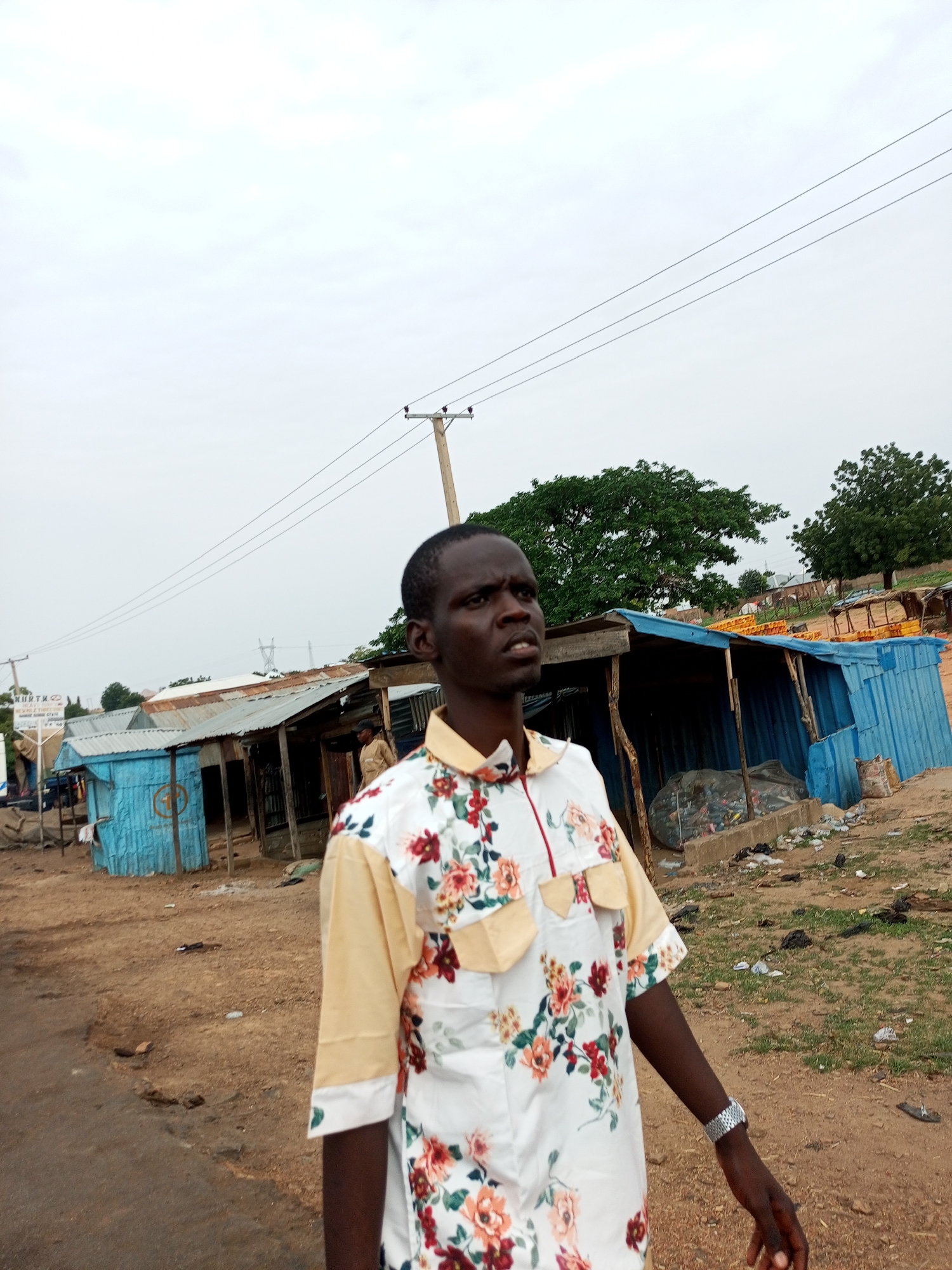 A young Black man, wearing a light-colored shirt with a floral pattern, stands in the foreground, looking off to the right. His left arm is extended slightly, with a watch visible on his wrist. Behind him, a dirt road leads to a row of small, weathered structures that appear to be market stalls or shops. The buildings have corrugated metal roofs and some are painted in shades of blue and gray. There are various items visible outside the stalls, including what looks like plastic bags and some yellow objects stacked on wooden pallets. A power line runs diagonally across the sky. In the distance, there are trees and some other structures, suggesting a rural or semi-urban setting. The sky is overcast, indicating it might be a cloudy or late afternoon. The scene appears to be in Gadao, Nigeria. A sign with illegible text is visible on the left side of the image.