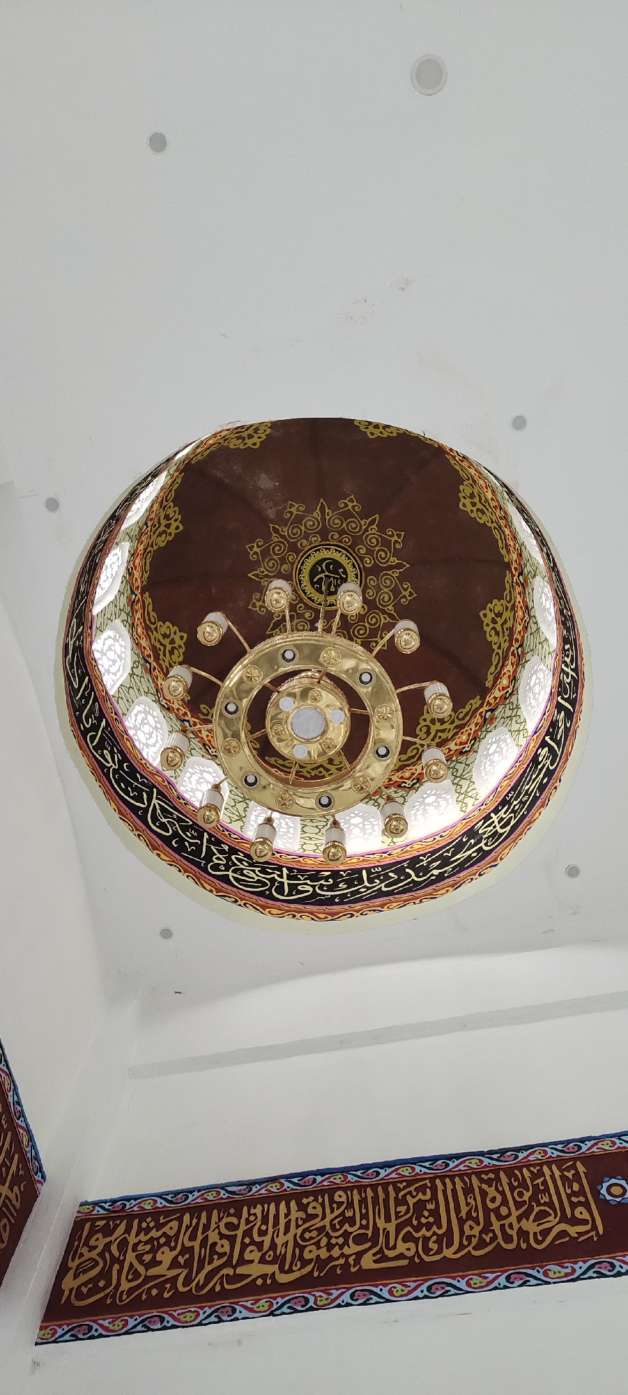 The image shows the interior of a building, likely a mosque, with a focus on its decorative ceiling and walls. A large, ornate golden chandelier hangs from the center of a domed ceiling. The dome is a deep brown color, adorned with intricate gold arabesque patterns and calligraphy. A band of white, perforated material, possibly a decorative screen, encircles the base of the dome. Below the dome, the walls are decorated with a horizontal band of calligraphy in gold lettering against a dark red background, bordered by colorful geometric patterns. The overall impression is one of grandeur and religious artistry. There is no visible activity or event taking place, and the lighting suggests an indoor environment, possibly during the day or under artificial light. The location is suggested to be a place of worship due to the presence of calligraphy and the architectural style, but the specific location is unknown.