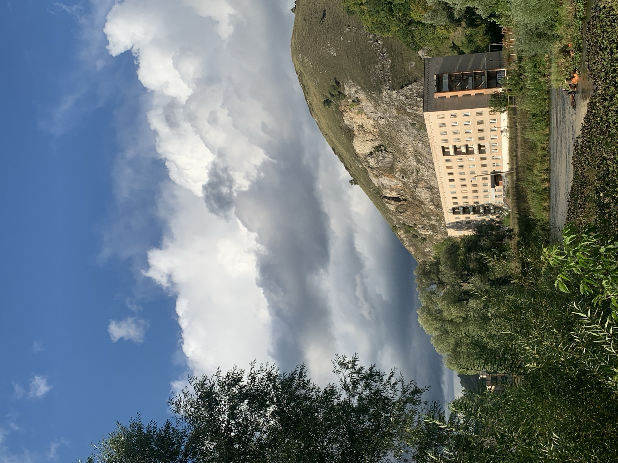 The image depicts a light brown, multi-story building situated near a rocky hillside and a body of water, likely a river. The building has numerous windows and balconies, and appears to be an apartment complex or similar structure. Dense green vegetation, including trees and bushes, surrounds the building and lines the riverbank. The sky is a mix of blue with scattered white and dark clouds, suggesting varied weather conditions. The scene is set against a backdrop of rolling hills, one of which is a prominent rocky incline. There is a single individual visible in the water near the riverbank, engaged in some activity with orange objects. The location is identified as the city of Asha, Russia.