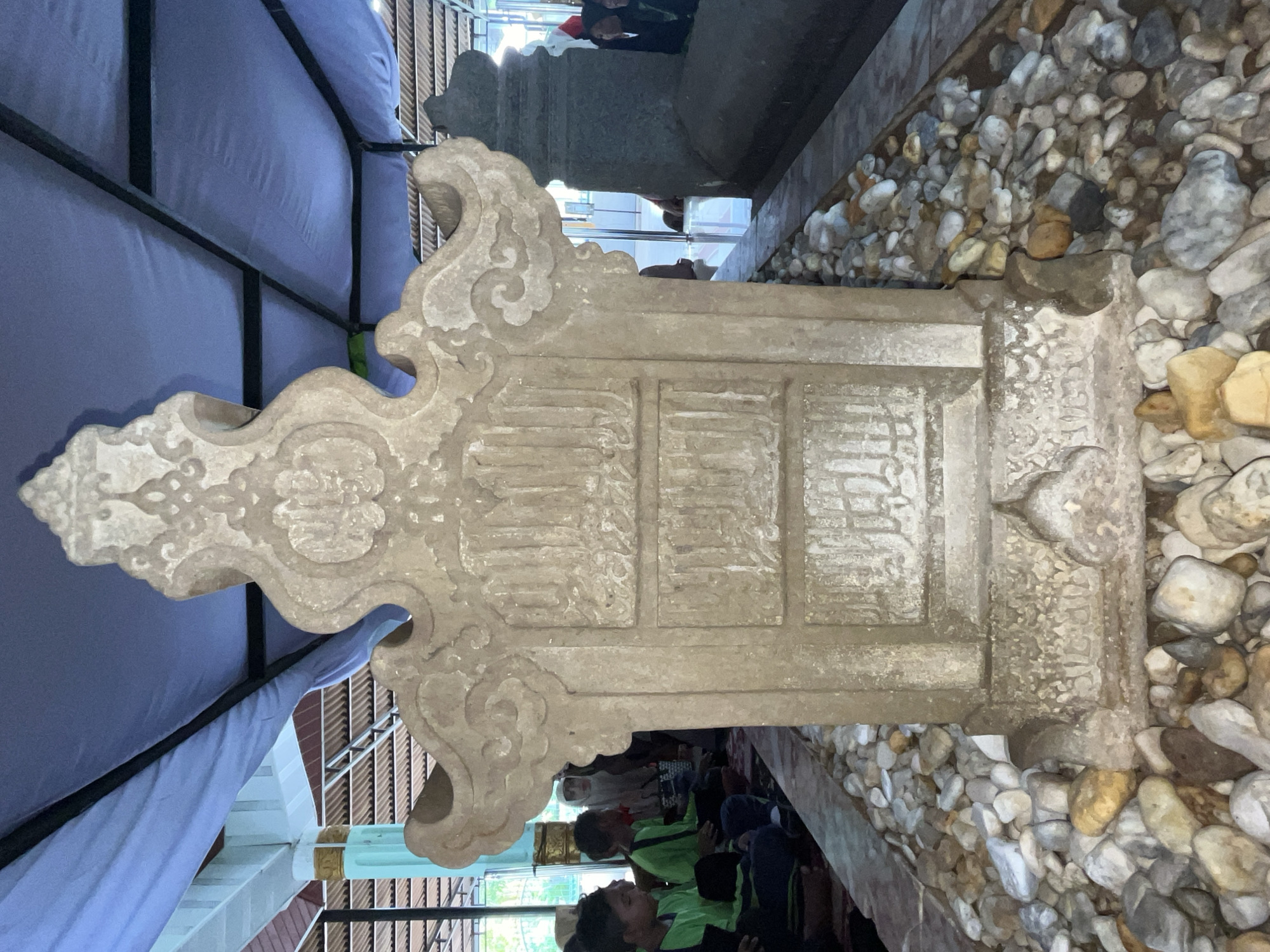 In Beuringen, Indonesia, a weathered stone tomb stands prominently in the foreground, surrounded by small, assorted pebbles. The tomb features ornate carvings and inscribed text, suggesting it is of historical or religious significance. Behind the main tomb, another, larger stone tomb is partially visible. Several people, including children and adults, are seated on the ground to the left of the central tomb, appearing to be in a solemn or contemplative state, possibly engaging in prayer or remembrance. They are partially covered by a blue tarp that extends overhead, providing shade. The environment appears to be an outdoor or semi-outdoor sacred site, possibly a cemetery or shrine, with architectural elements of a building visible in the background, including pillars and roofing structures. The lighting suggests it is daytime, with natural light filtering through the covered area.
