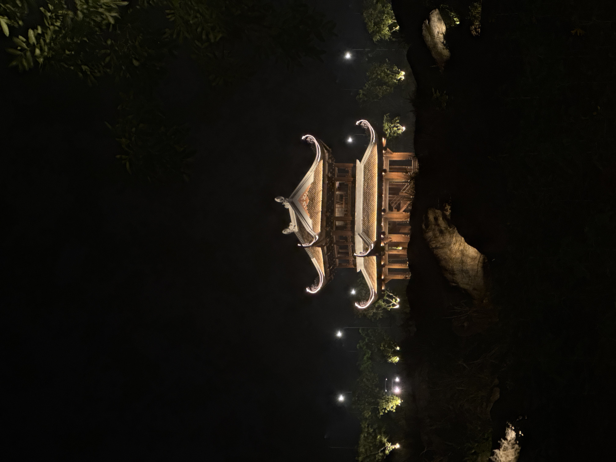 The image shows a pagoda at night in Tây Hoa Lư Ward, Vietnam. The pagoda is illuminated with warm, golden lights, highlighting its traditional architectural features, including tiered roofs with upturned eaves and intricate wooden detailing. It is situated on a slightly elevated, dark landscape with rocks and sparse vegetation visible in the foreground. In the background, several streetlights and scattered trees are visible, also illuminated by artificial lights, creating a softly lit, atmospheric scene against the dark night sky. A car with its headlights on is also visible in the distance.