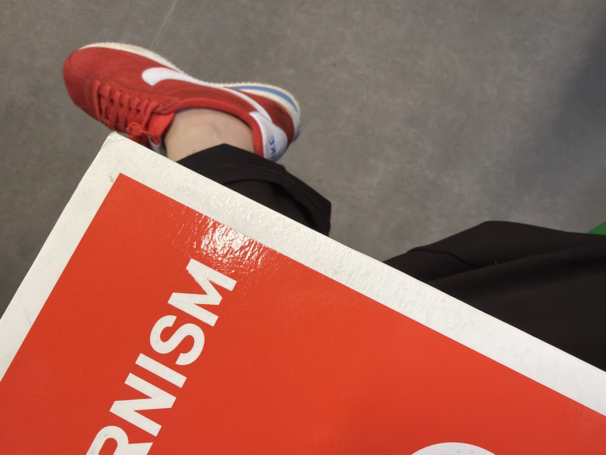 The image shows a person wearing red Nike sneakers, standing on a gray floor in Yerevan, Armenia. The person is holding an orange and white sign with the word "RNISM" printed on it. The person is wearing black pants. The background is a gray concrete floor.