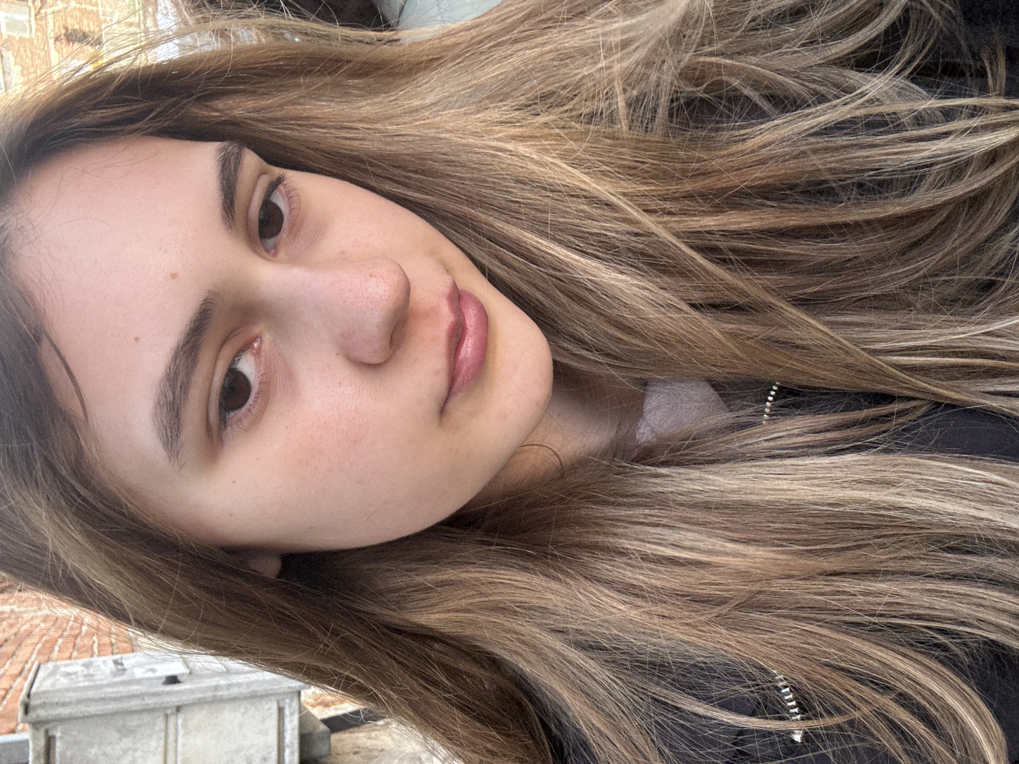 A close-up portrait of a young woman with long, light brown hair. Her skin is fair and she has brown eyes. She is wearing a dark jacket. The background is blurred, but it appears to be an outdoor setting, possibly a rooftop. A brick building and a metal box are visible. The location is likely Panagyurishte, Bulgaria.