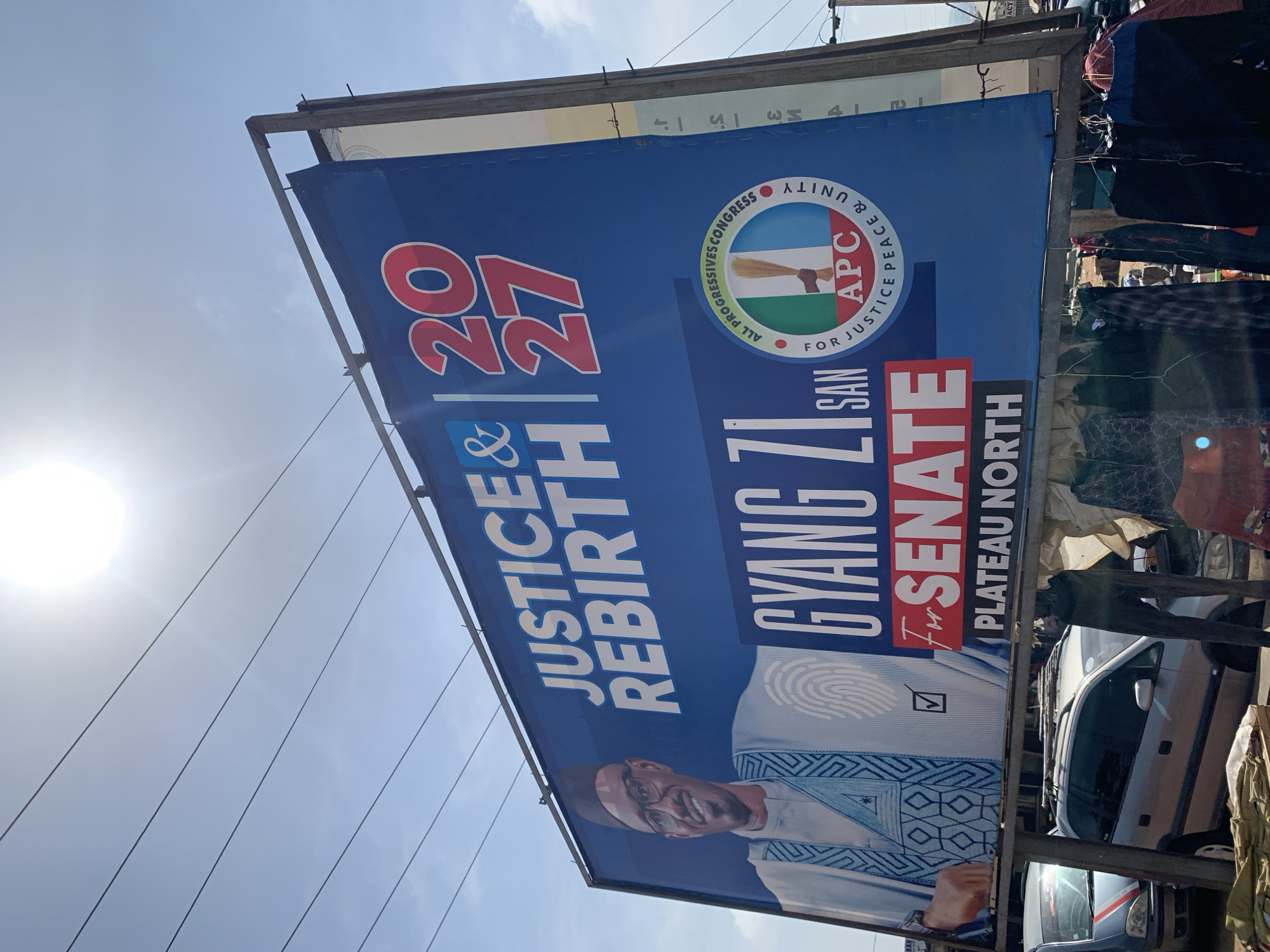 The image shows a political campaign billboard in Rafin Par, Nigeria. The billboard features Gyang Zi San, a candidate running for Senate in Plateau North. The billboard includes the candidate's photo, the slogan "Justice & Rebirth", the years "2027", and the APC party logo with the words "All Progressives Congress" and "For Justice Peace & Unity". Clothes are hanging from the metal framework of the billboard. There is a white car under the billboard and people behind it. The sky is visible in the background.