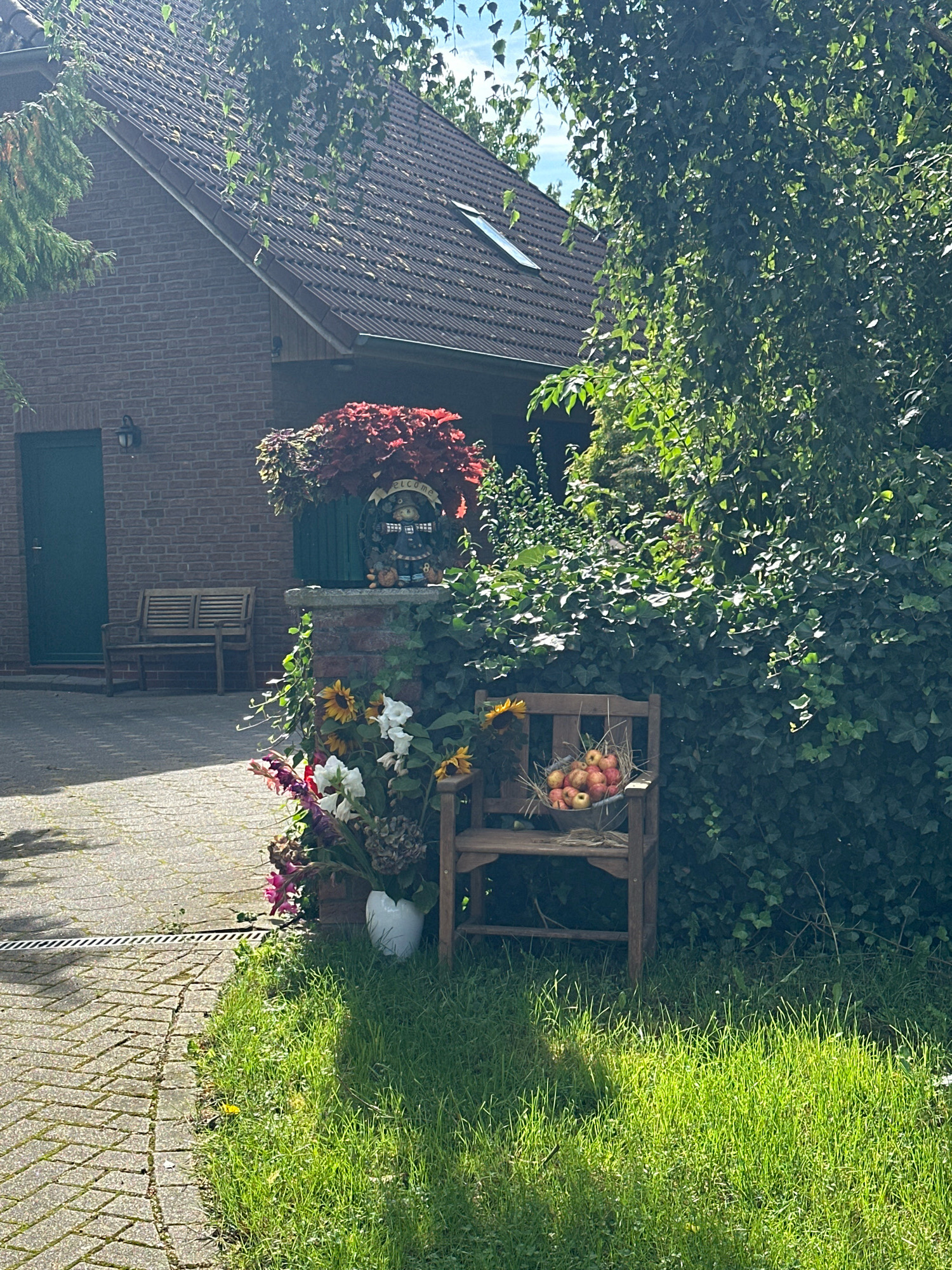 The image depicts a rustic, sun-drenched scene in front of a brick house, likely in the Samtgemeinde Thedinghausen, Germany. A wooden bench sits to the left of a dark green door. In the foreground, a small wooden chair is filled with apples. Adjacent to the chair, a white vase holds a bouquet of flowers, including sunflowers and gladioli. Lush green foliage frames the right side of the scene, creating a sense of natural abundance. The ground is a mix of paving stones and well-maintained grass. The sky above is partially obscured by the leaves of a large tree.