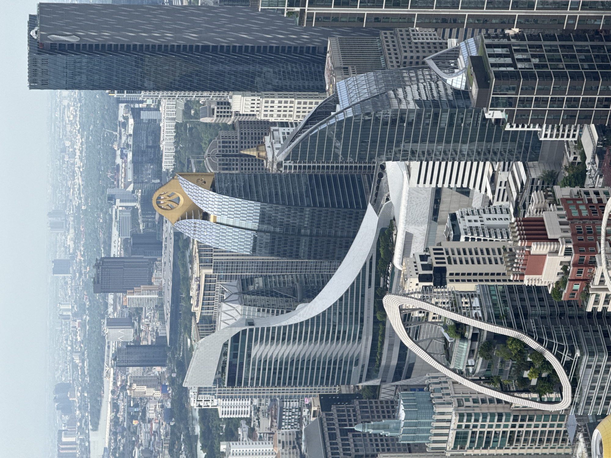 The image shows a high-angle, aerial view of the Bangkok skyline in Thailand. The shot is taken with a landscape orientation that is rotated 90 degrees. The modern urban landscape features various skyscrapers with unique architectural designs, including curved structures and glass facades. One notable feature is a prominent white loop-like structure. The city extends into the hazy distance.