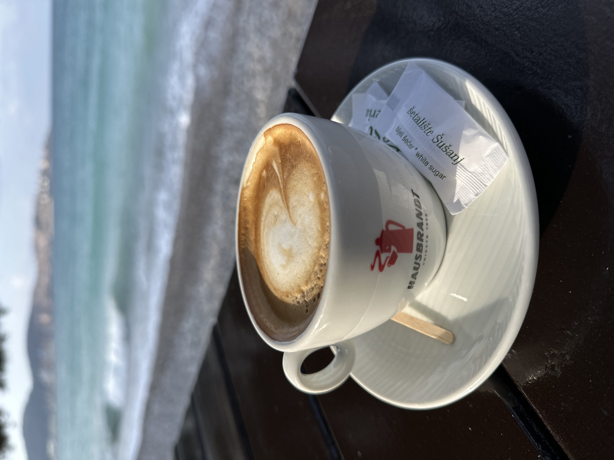 The image displays a close-up, slightly elevated perspective of a coffee cup and accessories situated on a dark table, with a coastal environment visible in the blurred background. The scene is located at Šetalište Šušanj (Šušanj Promenade) in Šušanj, Montenegro, confirmed by text on a sugar packet.
In the foreground, a white ceramic coffee cup, bearing the red "HAUSBRANDT TRIESTE 1892" logo featuring a stylized kettle, contains a light brown beverage topped with frothy milk, exhibiting a subtle latte art swirl pattern. The cup rests on a matching white saucer. Positioned on the saucer are a small, light-colored wooden stir stick and two white, rectangular sugar packets. The clearly visible sugar packet is printed with "Šetalište Šušanj" and "Bijeli šećer * White sugar." The cup, saucer, stir stick, and sugar packets are arranged on a dark, horizontally slatted table surface, which shows reflections of ambient light.
The background, out of focus, reveals a narrow, dark-toned beach with the turquoise and blue waters of the sea beyond it. Gentle waves are visible, breaking with white foam along the shoreline. In the distant background, blurred outlines of mountains or hills are discernible under a light sky. No individuals or specific actions are depicted in the frame.