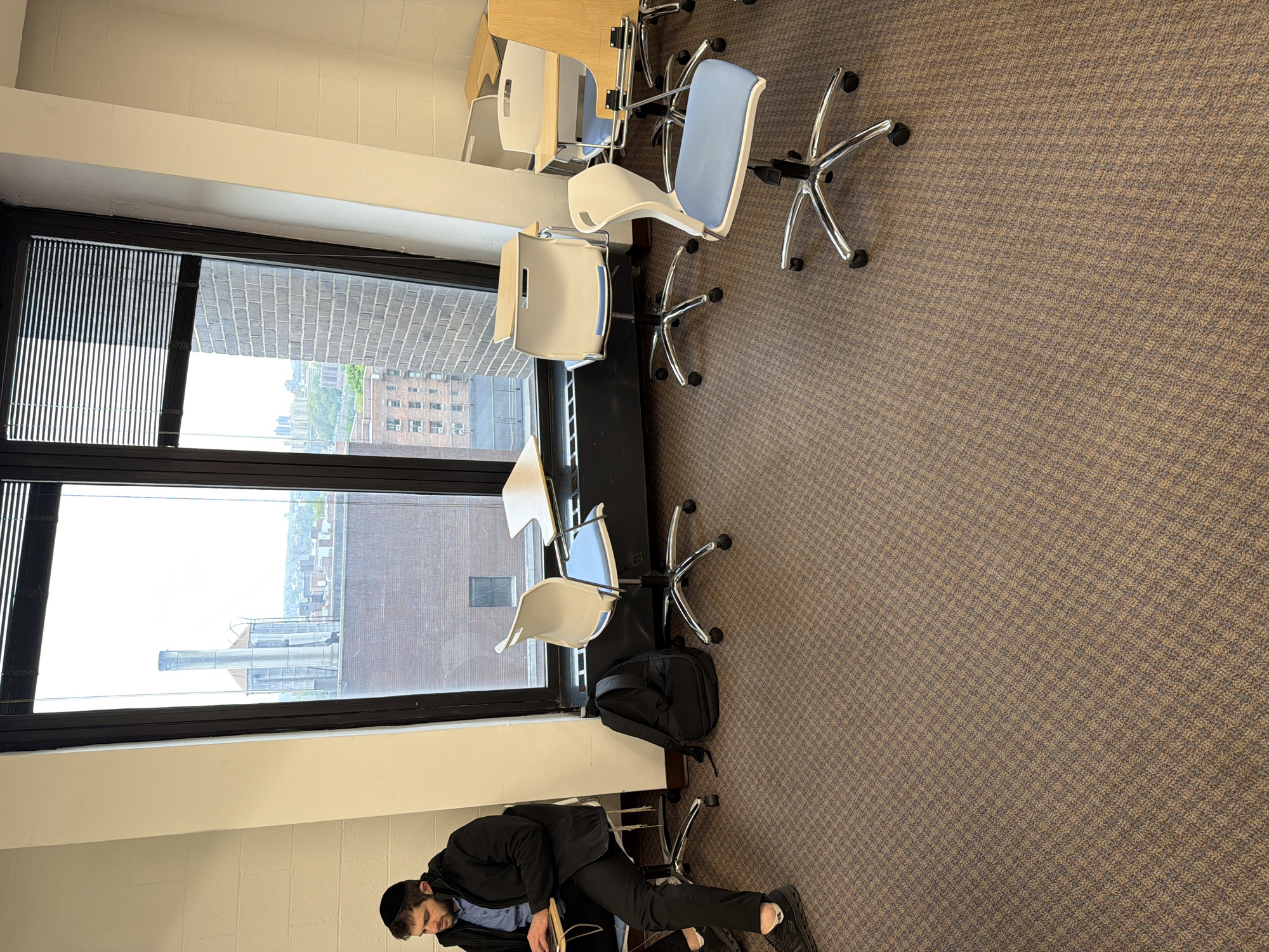The image depicts an indoor room, likely an office, classroom, or communal study area, distinguished by a large window spanning its length on the left side. The floor is covered with a patterned carpet in shades of brown and beige. Along the window, a series of light wood-colored desks are arranged, each paired with a modern office chair. These chairs feature chrome five-star bases with wheels, white plastic backs, and either white plastic or light blue upholstered seats.
Through the window, an urban landscape is visible under a bright, possibly overcast, daytime sky. The view includes several brick buildings, some with visible windows and air conditioning units, and a tall, industrial-style chimney or vent in the distance. Dark vertical blinds are partially drawn on the far left section of the window.
In the bottom right corner of the frame, a man is partially visible, appearing to be seated at one of the desks. He is wearing a dark jacket over a blue collared shirt, black pants, and a black yarmulke. His beard is also visible. A black backpack rests on the carpet next to a chair positioned further along the row of desks. The room appears orderly and suggests a quiet, functional environment. There is no discernible text within the image.