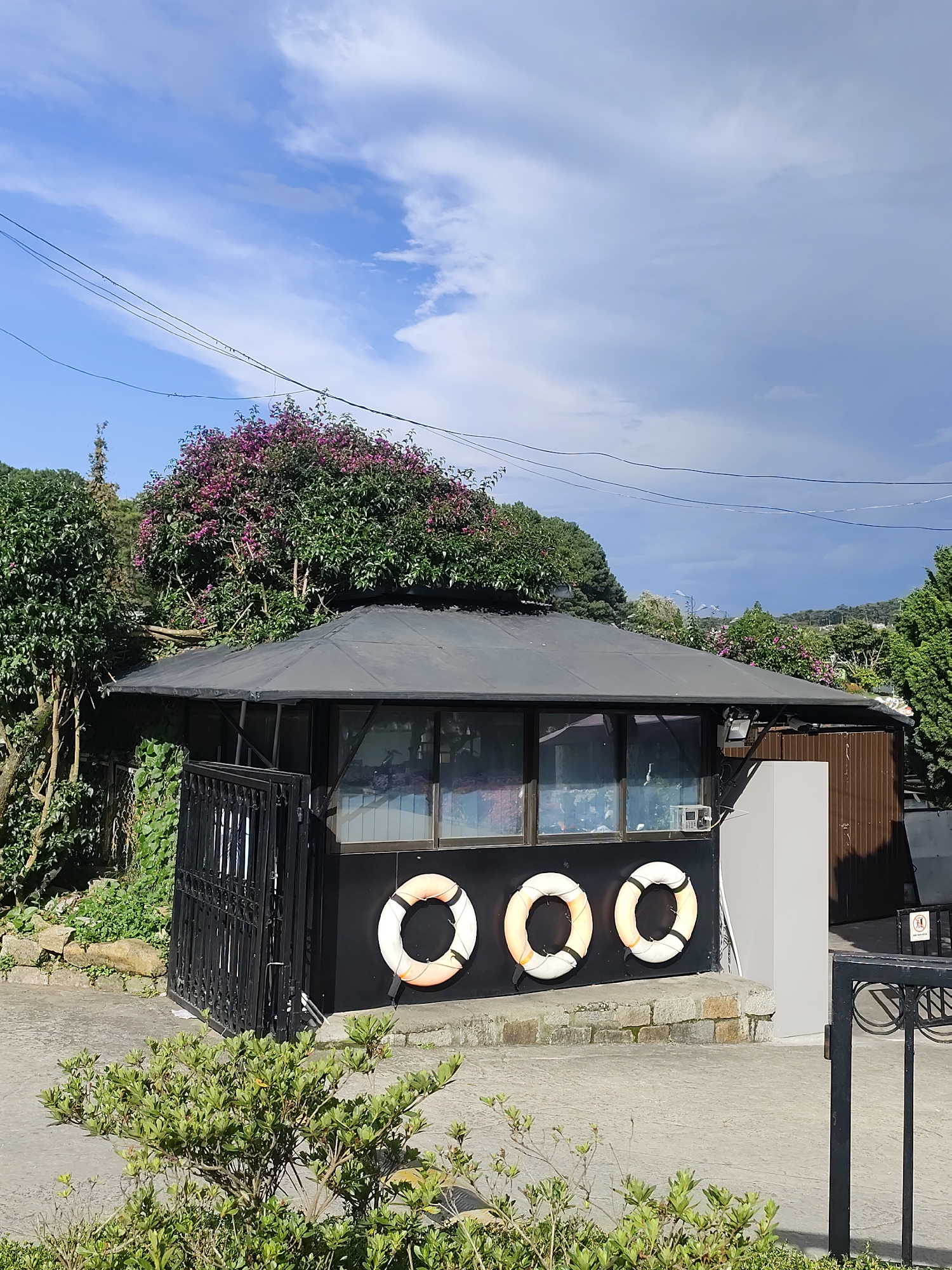 A small, dark-colored booth with a sloped, dark roof is situated outdoors. Three life preservers, white with orange stripes, are attached to the exterior wall of the booth. A black metal gate is to the left of the booth, and a light-colored rectangular structure is to the right. Green foliage and trees are visible in the background, with some trees bearing pink blossoms. The sky is blue with scattered white clouds. The scene appears to be in an urban or semi-urban outdoor setting, likely a park or recreational area in Da Lat, Vietnam, given the style of the booth and surrounding vegetation. There are no people or visible actions in the image.