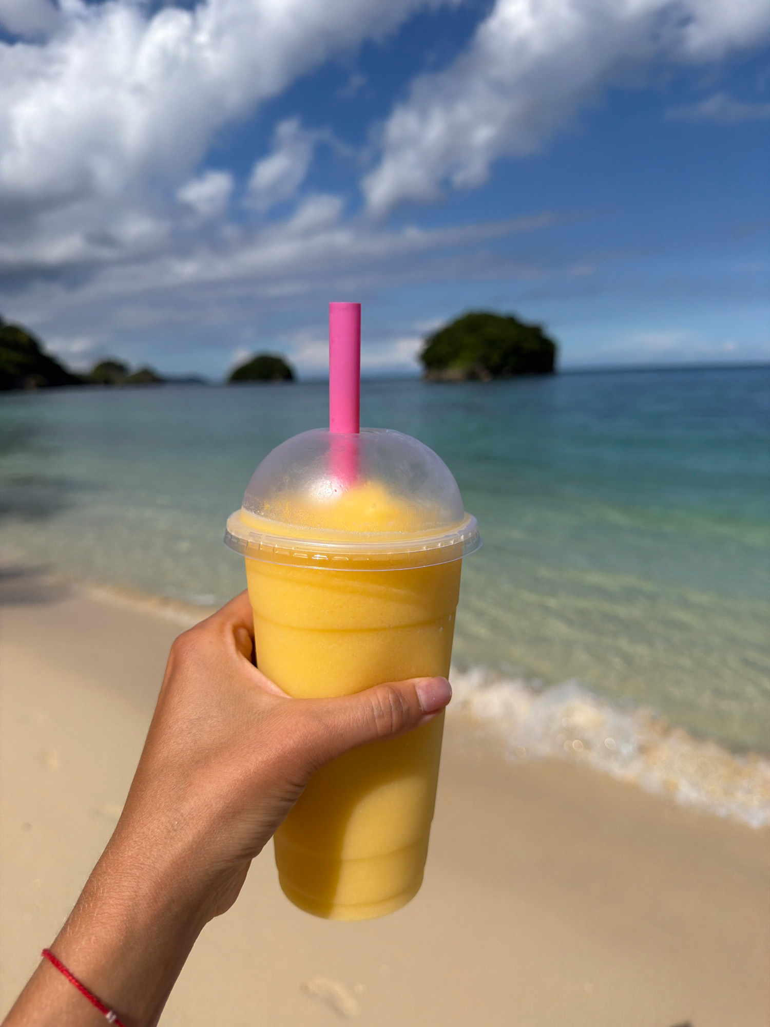 A hand holds a clear plastic cup filled with a yellow frozen beverage and a pink straw. The cup is in the foreground, positioned on the right side of the image. The background shows a sandy beach, turquoise ocean water, and a few small, tree-covered islands under a cloudy blue sky. The scene appears to be located in the city of Malay, Philippines, likely on one of its numerous beautiful beaches.