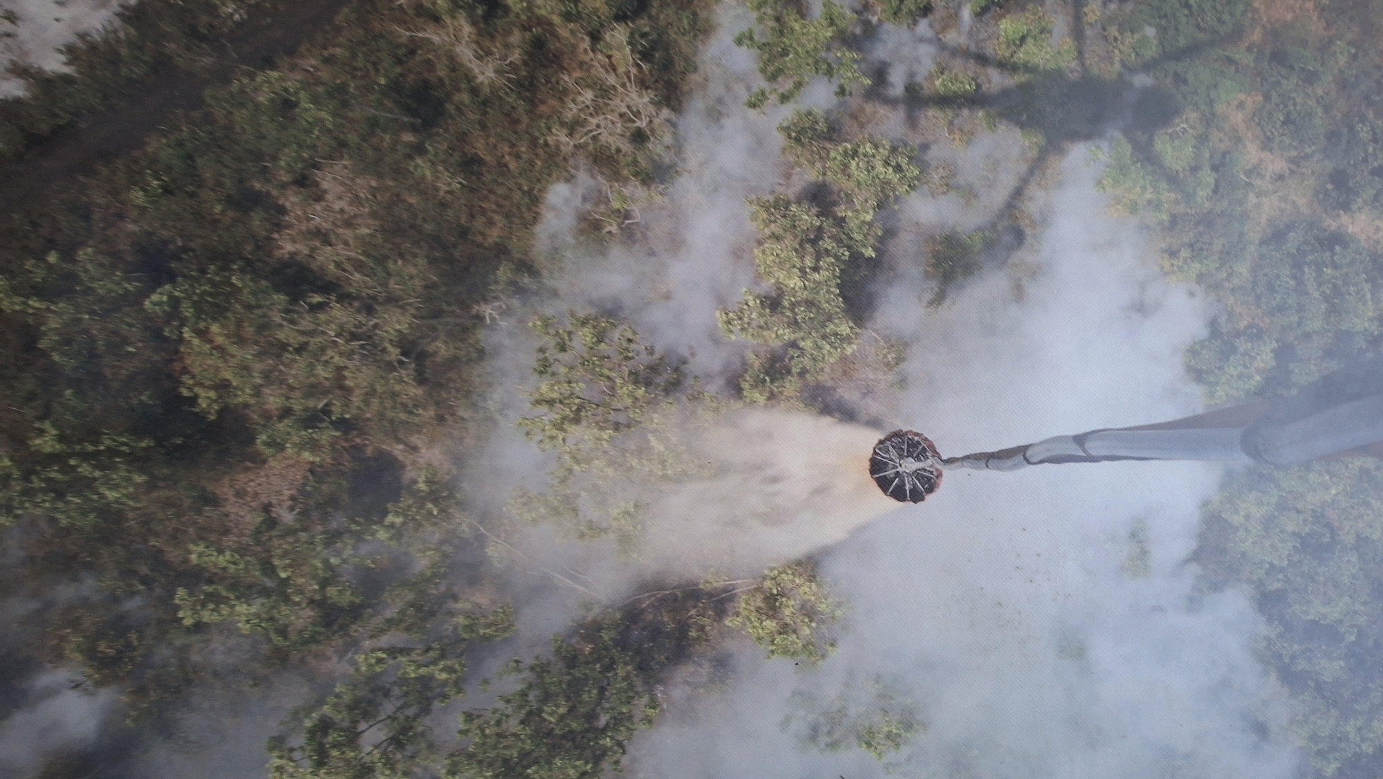An aerial, top-down view captures a firefighting operation in a densely vegetated area, likely a forest or park within Palembang, Indonesia. A large, circular water bucket, suspended from a cable, is actively dropping water onto a patch of smoldering vegetation, indicated by a visible plume of grey smoke. The bucket appears to be part of a heavy-lift helicopter or a similar aerial firefighting system. The surrounding environment consists of numerous trees with green and brown foliage, interspersed with areas of dry, brown undergrowth, suggesting a potential wildfire or controlled burn. The smoke obscures portions of the landscape and the firefighting equipment. The lighting suggests it is daytime.