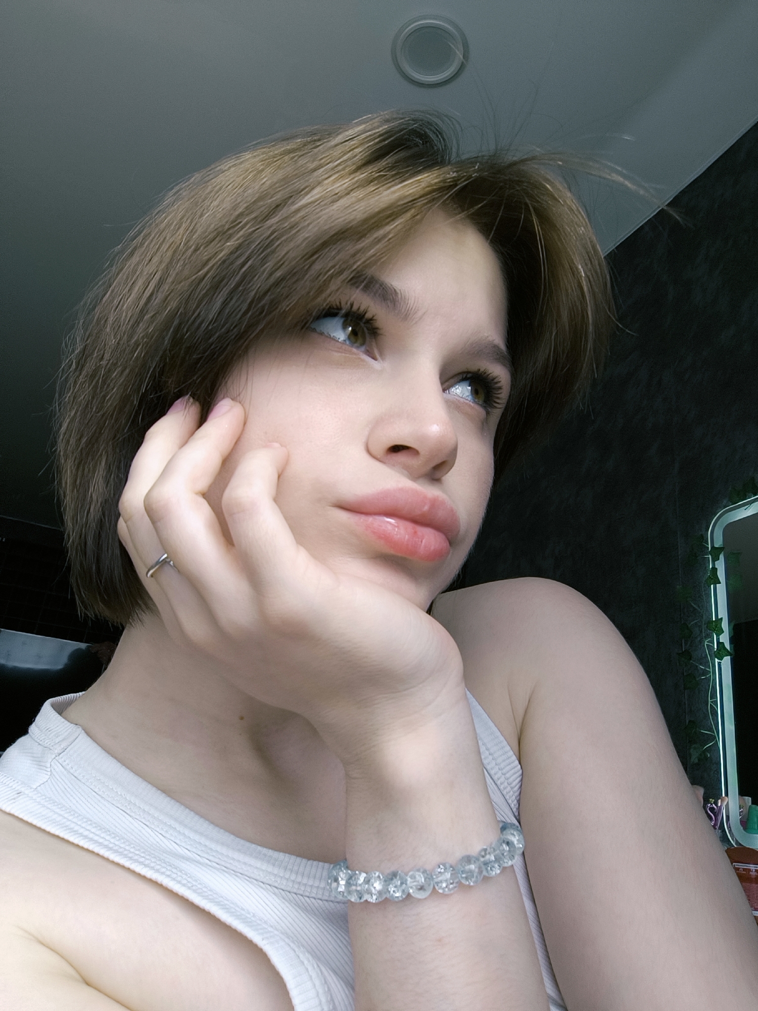 A young woman with short, brown hair is posed in a thoughtful or contemplative manner, resting her face on her hand. She is wearing a white tank top. Her skin appears light-toned. A silver ring is visible on her finger. A light blue beaded bracelet is visible on her wrist. The background shows a dark wall and a glimpse of a mirror with what appears to be artificial ivy. Above, a circular ceiling fixture is visible. There is nothing in this image to indicate the location in the city of Chaikovsky, Russia.
