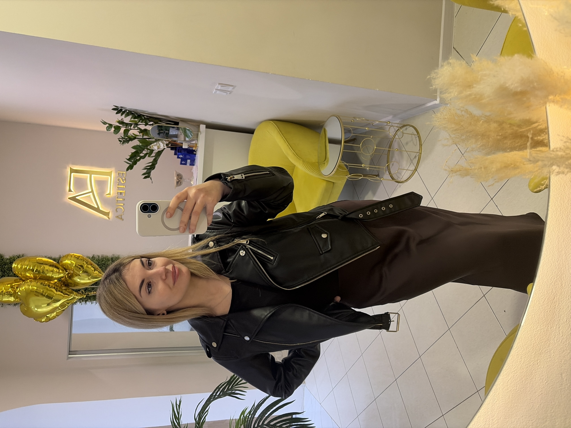 A woman is taking a selfie in a mirror within a commercial establishment, likely a salon or beauty studio. The location is potentially in Budva, Montenegro, indicated by the establishment's illuminated sign reading "FA ESTETICA."
The woman is wearing a black leather jacket over a dark dress or top. She is holding a light-colored smartphone in her right hand to capture the image. Her hair is blonde.
The environment includes elements such as a yellow chair, a gold-colored side table, a plant arrangement, and decorative elements, including some pampas grass. The setting appears well-lit and modern.