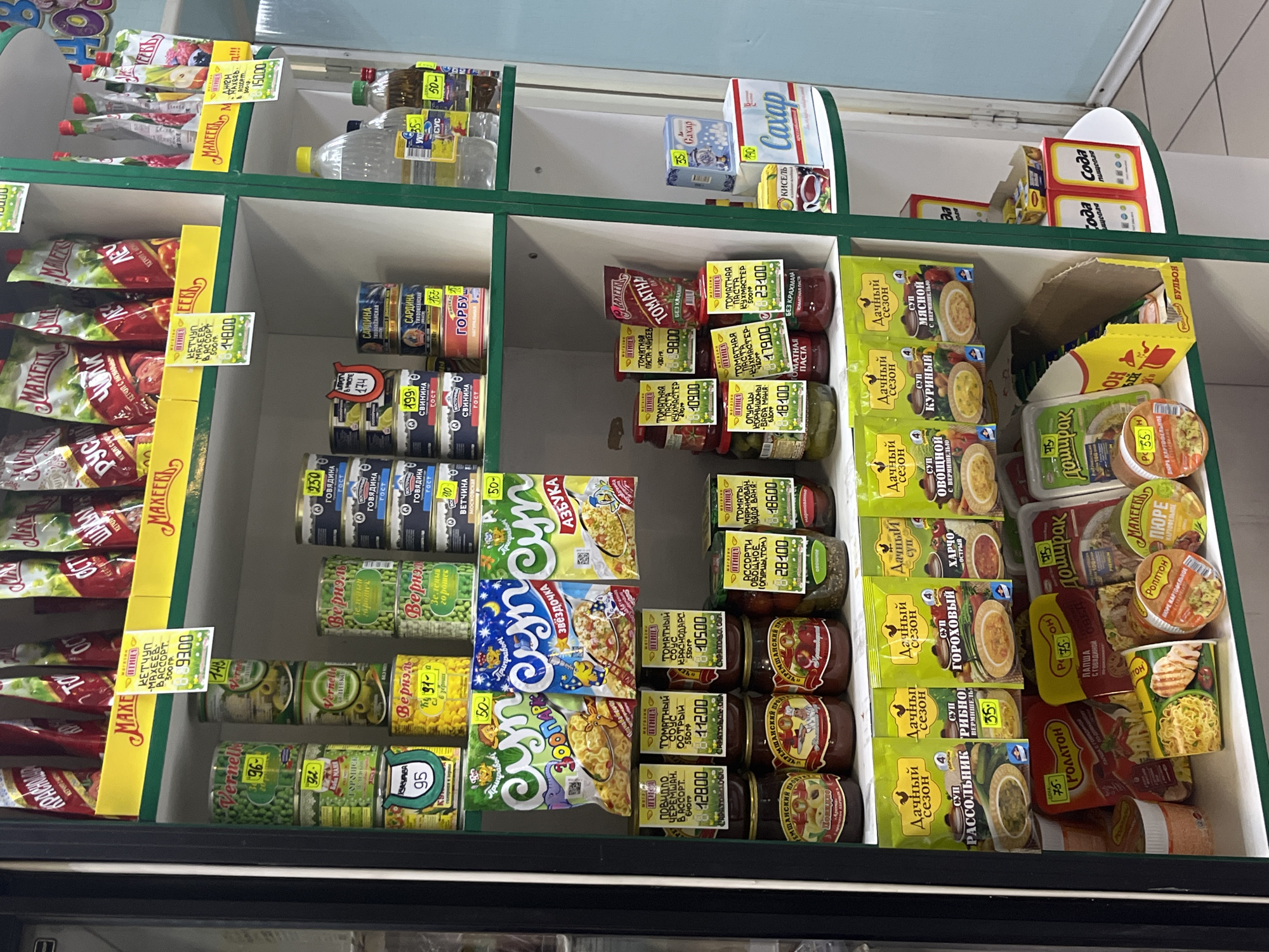 The image shows the inside of a refrigerated display case, likely in a grocery store in Фоки, Russia, filled with various food products. The case is divided into shelves.
On the top shelf, there are bottles of liquid (possibly vinegar or oil), small boxes of "Gaxap", and other packaged goods.
The middle shelves contain canned goods such as fish (sardines, salmon), vegetables (peas), and jars of tomato paste, pickled cucumbers, and other preserved goods. There are also packages of instant noodles ("Zvezdochka," "Azbuka") and instant soups ("Dachniy Sezon"). Many products have price tags attached to them.
The bottom shelves feature more instant soups, mashed potato flakes, and preserved vegetables. Brands visible include "Rollton", "Doshirak", "Maxeev", and "Vernelle".
The case is illuminated and appears to be well-stocked. The products are neatly arranged, and price tags are visible on many items. The overall impression is of a typical grocery store refrigerator section in Russia.