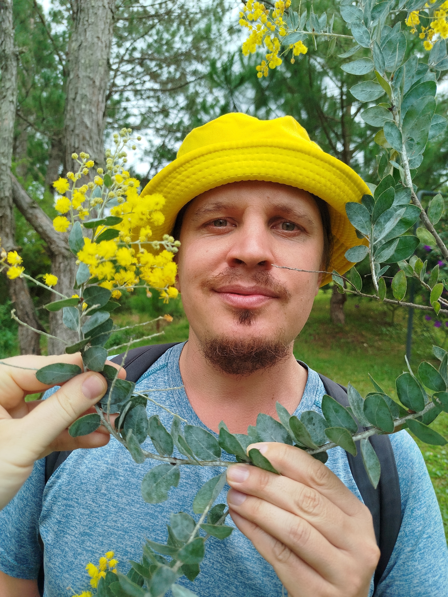 In Lâm Viên - Đà Lạt, Vietnam, a man wearing a yellow bucket hat and a blue t-shirt is holding a sprig of yellow flowers and green leaves in front of his face, obscuring the left side of his face. The man has a goatee and a mustache. He is outdoors, with trees and greenery in the background. The image is taken from the chest up.