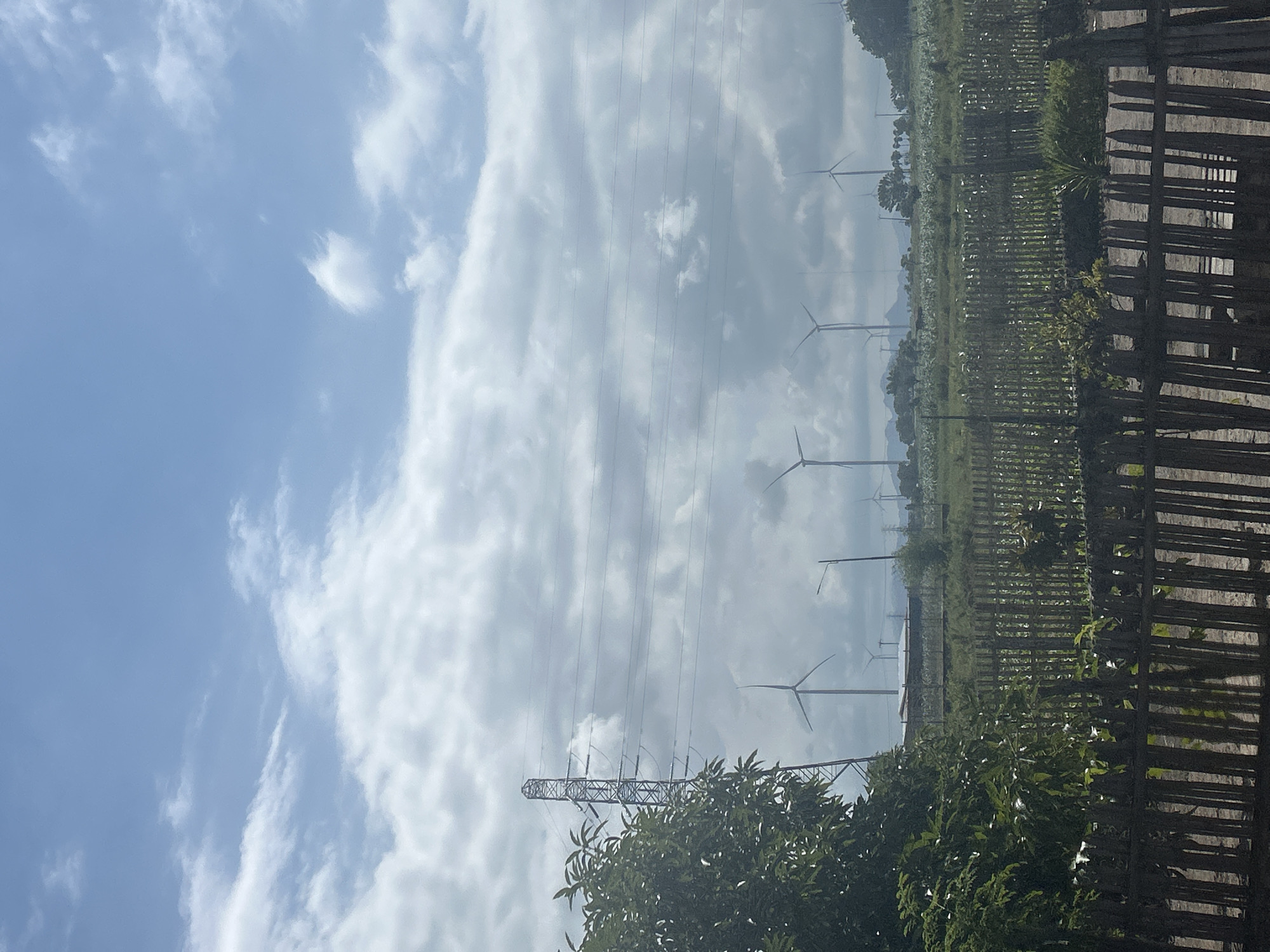 The image depicts a rural landscape in Jeneponto, Indonesia, characterized by a vast expanse of sky with scattered white clouds above a field of wind turbines. A prominent transmission tower stands to the left, its metallic structure silhouetted against the sky. In the foreground, a rustic wooden fence runs horizontally across the frame, separating the viewer from a lush green area with trees and vegetation. The background reveals a line of wind turbines, their blades visible against the horizon, suggesting a renewable energy installation. The overall scene is serene and natural, with a focus on the interplay between man-made structures and the environment.
