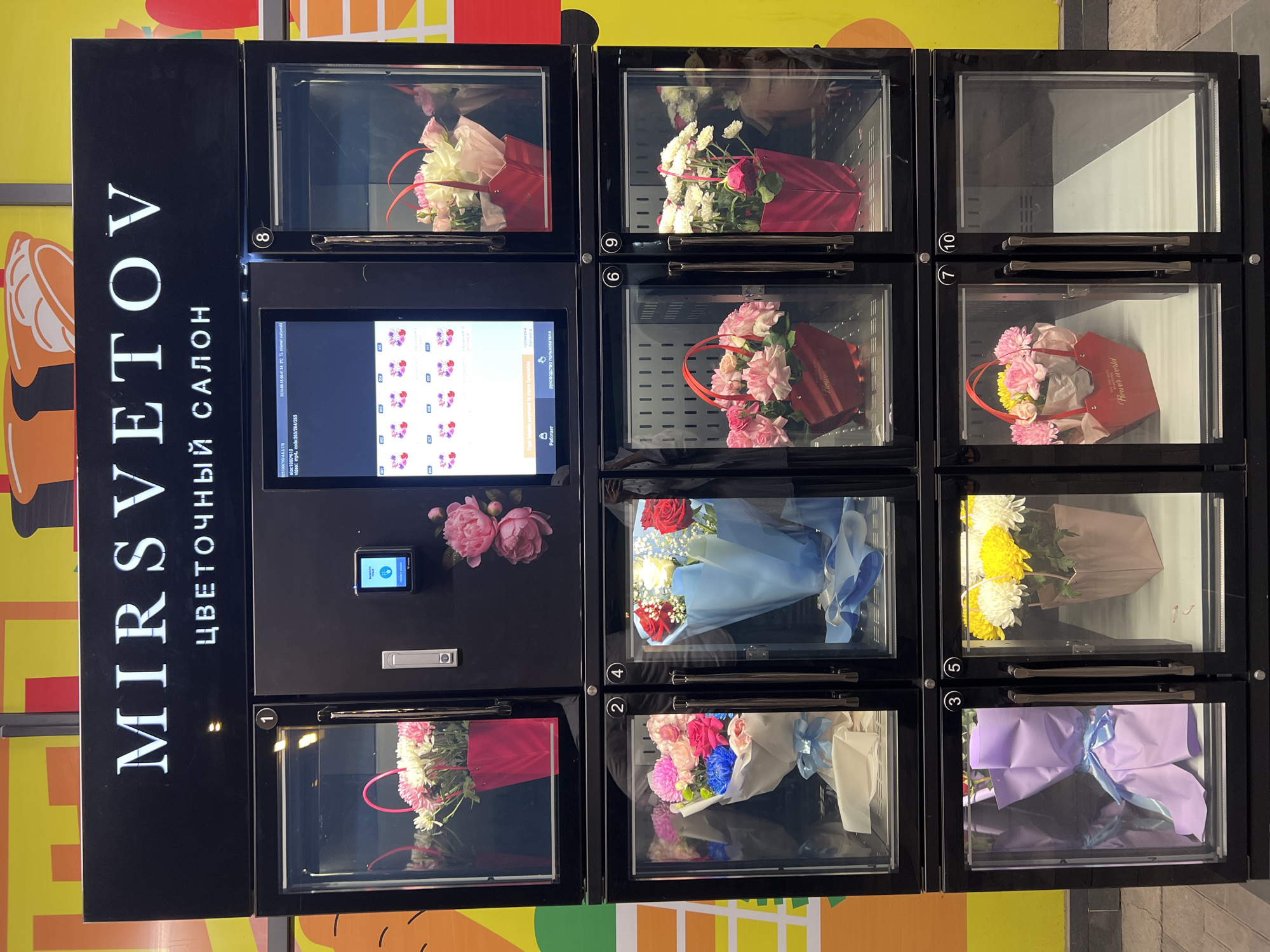The image shows a vertical flower vending machine with multiple glass-fronted compartments, each displaying a bouquet or flower arrangement. The machine has a black exterior with a prominent white illuminated sign at the top reading "MIRSVETOV" in large letters, followed by "ЦВЕТОЧНЫЙ САЛОН" (Flower Salon) in smaller letters below. The compartments are numbered from 1 to 10, though not all are visible or contain flowers.
Compartment 1, on the top left, contains a pink bag filled with white and pink flowers. Compartment 8, on the top right, has a red bag with a bouquet of white and pink flowers.
The middle row shows compartment 2 on the left with a bouquet of pink, blue, and white flowers wrapped in white paper and tied with a blue ribbon. Compartment 4, to its right, displays a bouquet of red and white roses wrapped in blue paper and tied with a blue ribbon. Compartment 6, on the bottom left, contains a red bag with pink and yellow flowers, featuring the "Flower World" logo. Compartment 9, on the bottom right, has a red bag with a bouquet of white and pink flowers. Compartment 3, on the bottom left, shows a bouquet wrapped in light purple paper and tied with a blue ribbon. Compartment 5, to its right, displays a pink bag with white and yellow chrysanthemums. Compartments 7 and 10 are empty or not clearly visible.
A digital screen is visible in the upper center of the machine, displaying flower options with numbers like "007," "008," "009," and "010." Below this screen are buttons for "Работая" (Working) and "Руководство пользователя" (User Manual).
The background of the image is a brightly colored wall, likely part of a building or advertisement, with yellow and orange sections. The vending machine is situated outdoors, with a paved ground visible in the foreground. There are no people clearly visible in the image. The image is focused on the flower vending machine itself.