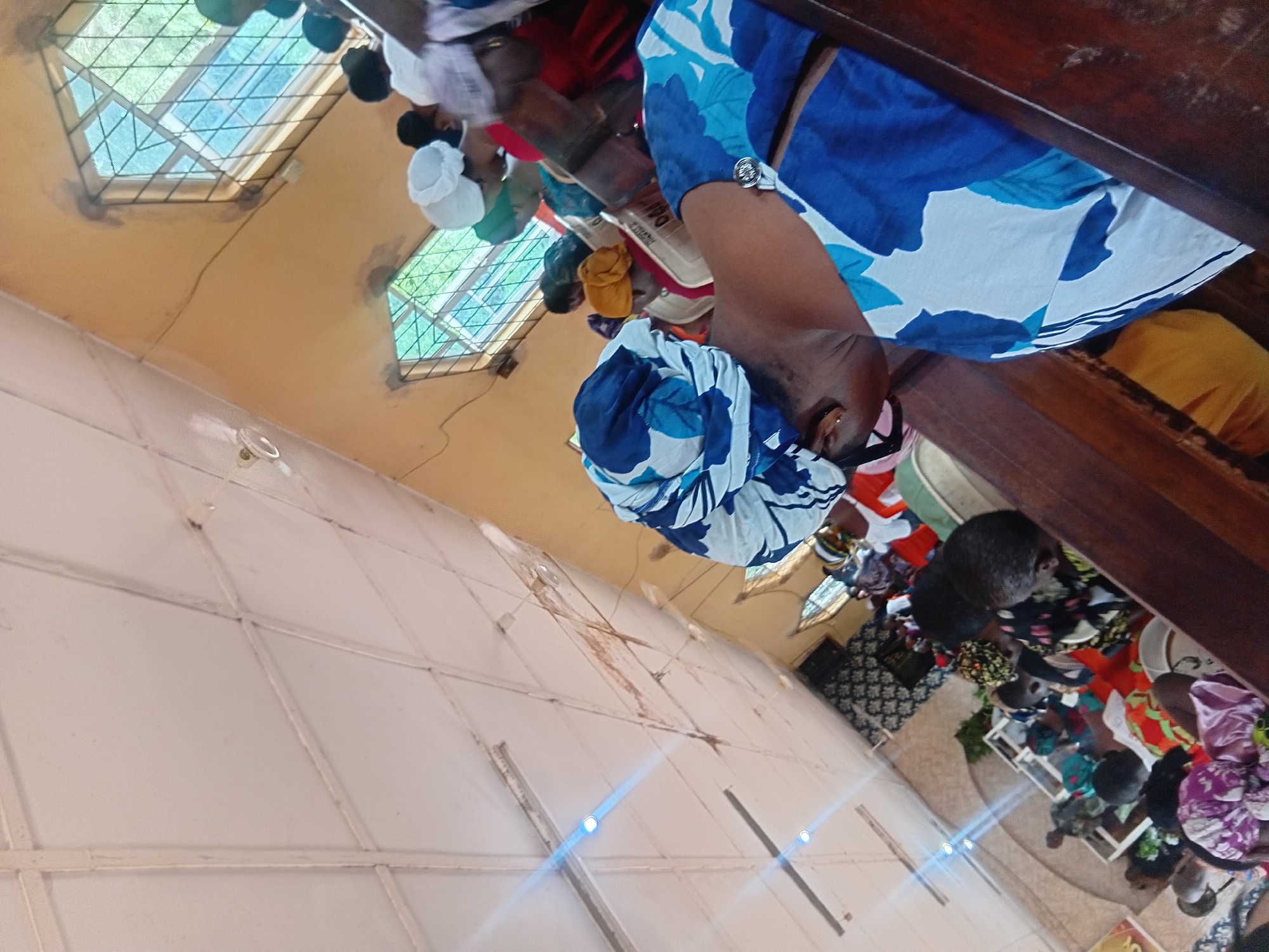 The image captures an indoor scene, likely a place of worship or a community gathering, in Ejule, Nigeria. The perspective is from a high angle, looking down into a seated congregation. The ceiling is a white, segmented paneling with some visible signs of wear and tear, along with lighting fixtures and electrical conduits. The walls appear to be painted a light ochre color. Two diamond-shaped windows with green-tinted glass are visible, offering a glimpse of foliage outside.
The foreground is dominated by the back of a person wearing a vibrant blue and white patterned garment and headscarf. To their right, another individual is visible, also wearing a head covering. The seating appears to be wooden benches or chairs.
Further into the scene, numerous people are seated, many of them women wearing colorful headwraps in various shades, including blue, white, yellow, and purple. Some individuals are leaning forward, suggesting prayer or attentiveness. The overall atmosphere appears to be one of a communal gathering, possibly a religious service given the attire and the setting. The lighting in the room is a mix of natural light from the windows and artificial lighting from the ceiling fixtures, creating a warm and slightly diffused illumination. The image is taken in portrait orientation.