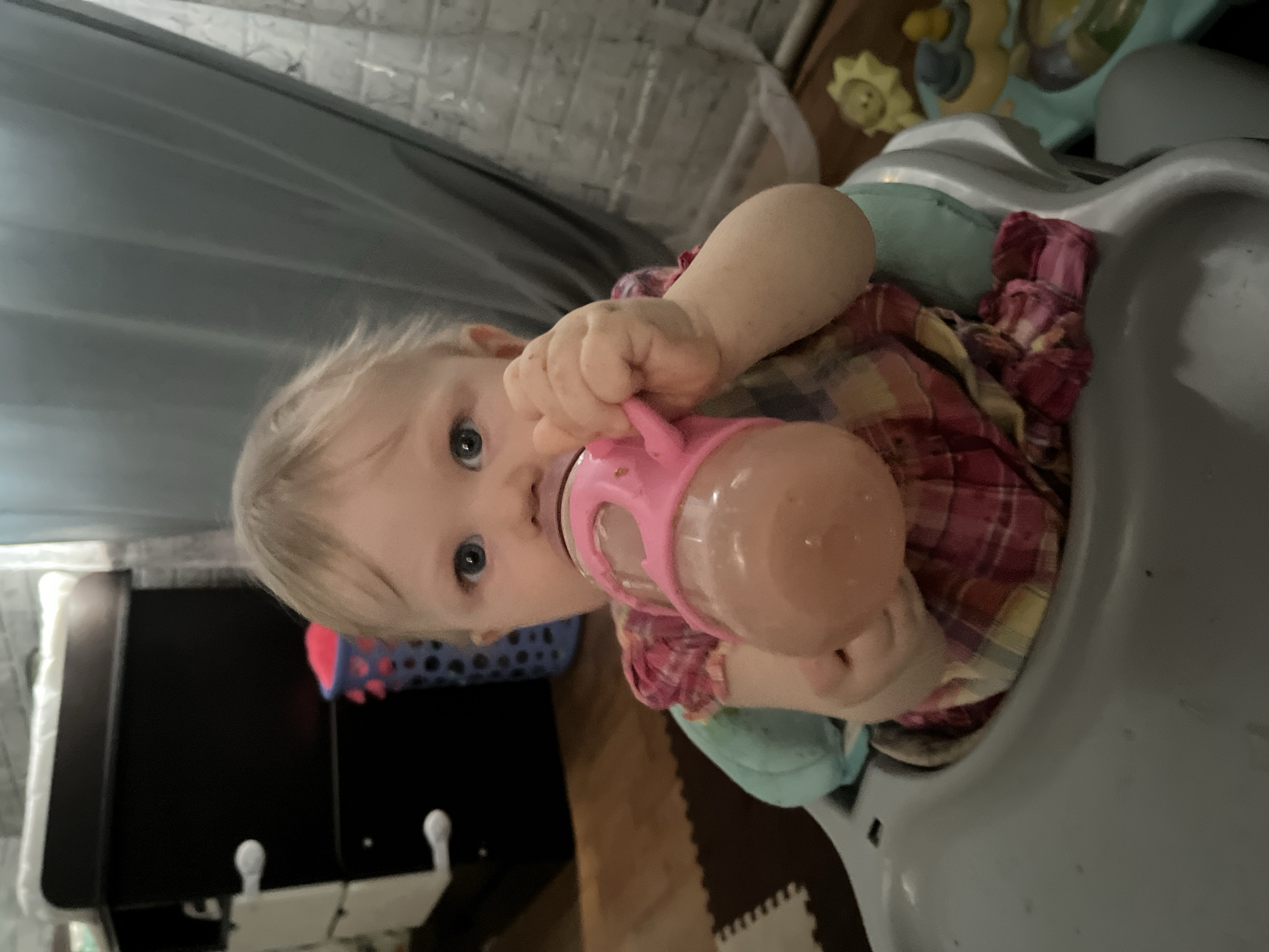 A baby with blonde hair and blue eyes is sitting in a gray high chair, holding a pink bottle to its mouth. The baby is wearing a plaid shirt. In the background, there is a blue curtain, a black cabinet, and a child's toy. The location is not Vesyoloye, Russia.
