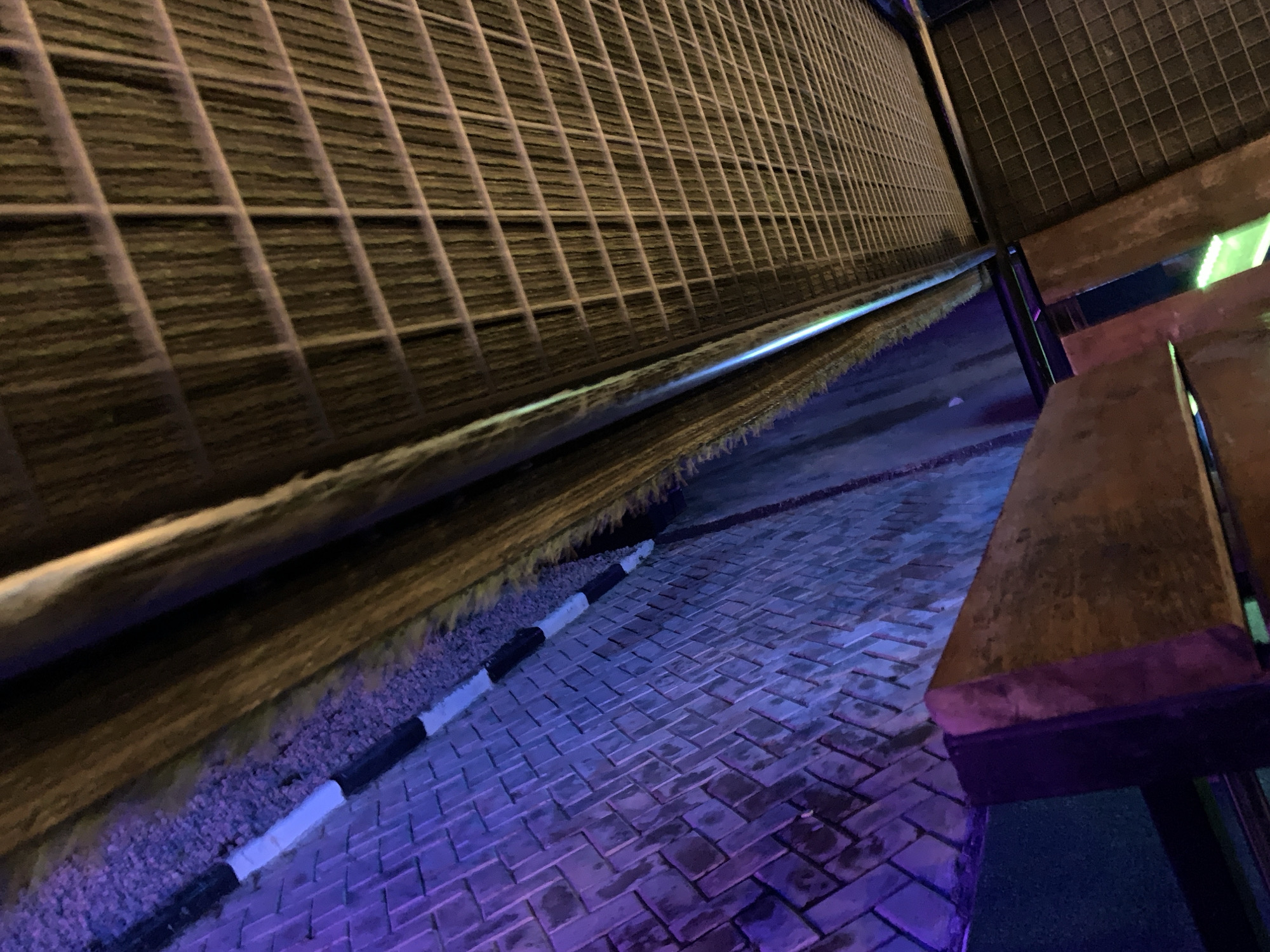 The image shows an outdoor scene at night, likely in Gura Top, Nigeria, with a focus on patterned brickwork and textured surfaces. In the foreground, a path of dark, rectangular bricks is illuminated with purple and blue light, suggesting artificial lighting. A curb with alternating black and white markings runs along the left edge of the path, separating it from a gravelly area and a dark, textured wall. This wall, possibly made of woven material or dense vegetation, has horizontal lines and is lit with a warm, golden-yellow light. Above this wall, a metal grid structure is visible, casting angular shadows and reflecting some light. In the upper right portion of the image, wooden benches are present, one of which is partially in shadow, while another is illuminated with a bright green light. The overall impression is of an urban or semi-urban outdoor space, possibly a seating area or walkway, at night, with deliberate use of colored lighting to create a specific ambiance. There are no people or animals visible in the image.