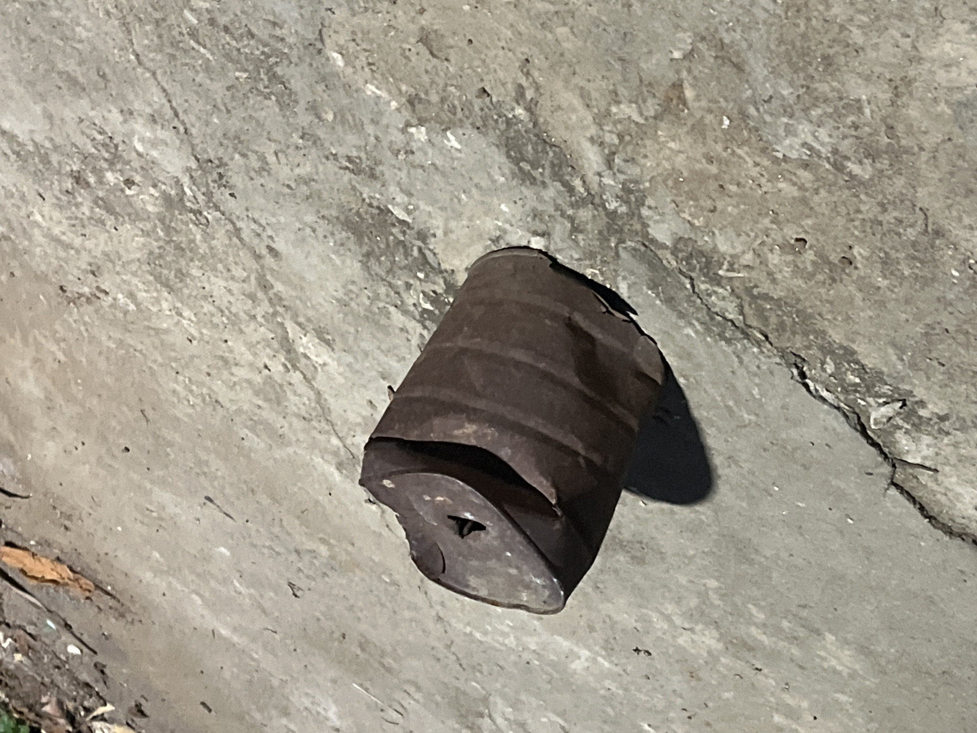 The image depicts a rusty, dented tin can lying on a cracked concrete surface. The can is positioned horizontally, with its opening facing left. The concrete ground shows signs of wear and tear, with visible cracks and small debris scattered on it. A shadow is cast by the can to its right, suggesting the presence of a light source from the upper left. The overall scene appears to be outdoors, possibly in an urban or industrial setting. No people are visible in the image. The location in the city of Santa Ana, Venezuela, is not identifiable from the image content.