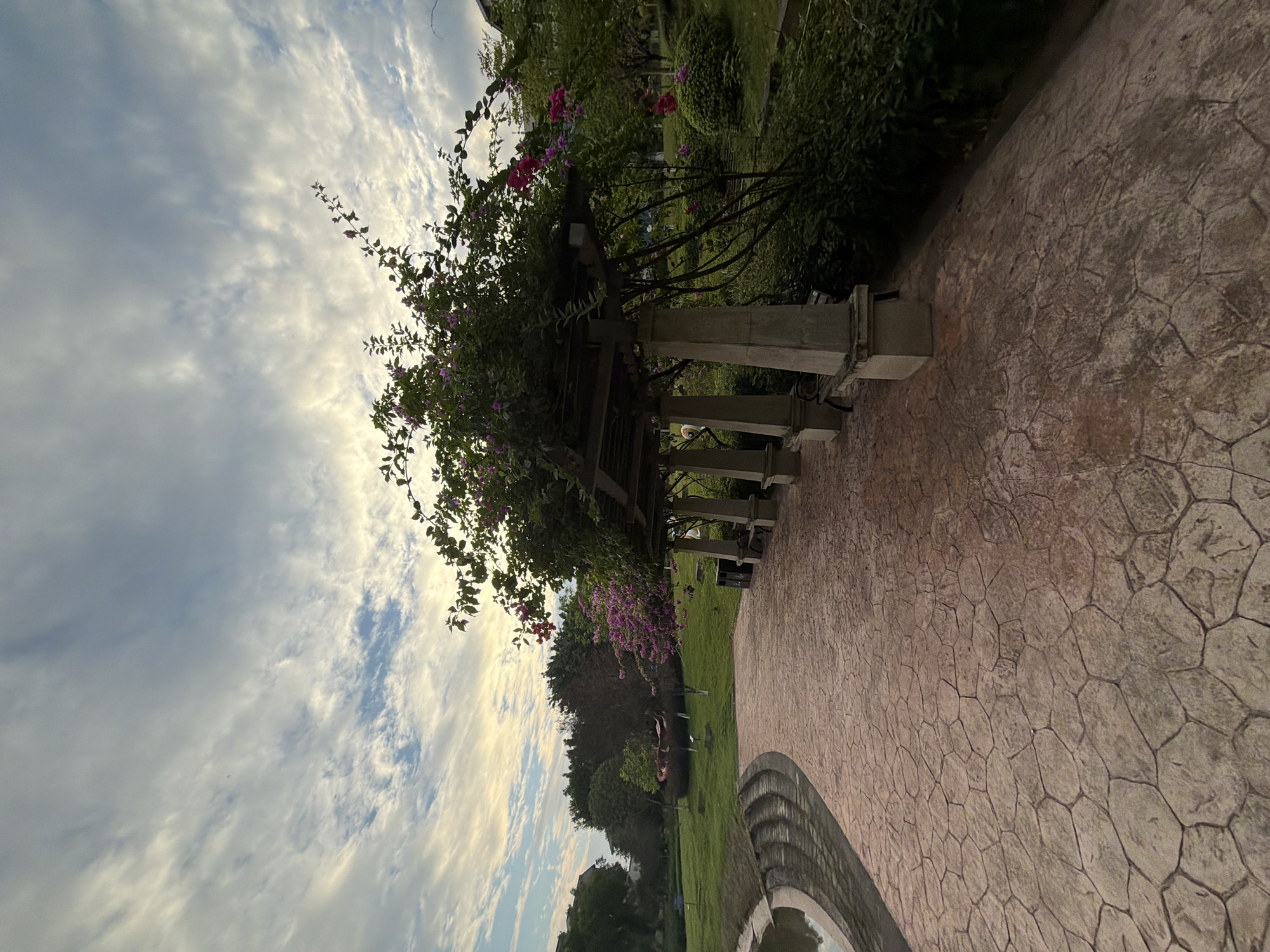 A scenic park pathway curves alongside a pond, its surface paved with hexagonal-shaped stones. A wooden pergola, supported by concrete pillars, offers shade and is adorned with vibrant pink and purple bougainvillea flowers. Benches are placed along the pergola, inviting rest. Beyond the path and pergola lies a lush green lawn dotted with trees, and in the distance, buildings are visible, suggesting an urban setting. The sky above is a canvas of dramatic clouds, with hints of sunlight breaking through. The scene captures a tranquil moment in a park, possibly in the city of Jiangjin, China, a place known for its natural beauty and public green spaces.