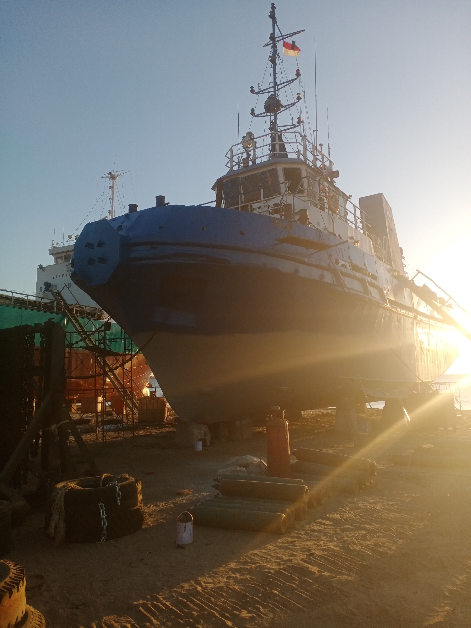 The image depicts a large, blue and gray vessel undergoing repair or maintenance in a dry dock or shipyard. The vessel, which appears to be a tugboat or a similar workboat, is elevated on supports. Its bow is prominently featured in the foreground, showing a distinct, angular design. The superstructure of the vessel is visible behind the bow, with various antennas and a flag flying from the mast.

The environment is sandy, suggesting a coastal location. Several objects are scattered around the dry dock, including stacks of cylindrical pipes, a red gas cylinder, and old tires wrapped with chains. In the background, other vessels and industrial structures are partially visible. The scene is illuminated by the warm light of a setting or rising sun, casting long shadows across the ground and creating lens flare effects. The sky is clear and blue.

The image does not contain any people. The overall impression is one of industrial activity and maritime operations. There is no indication of the city of Siddo, Indonesia, within the image itself; this information is external to the visual content.