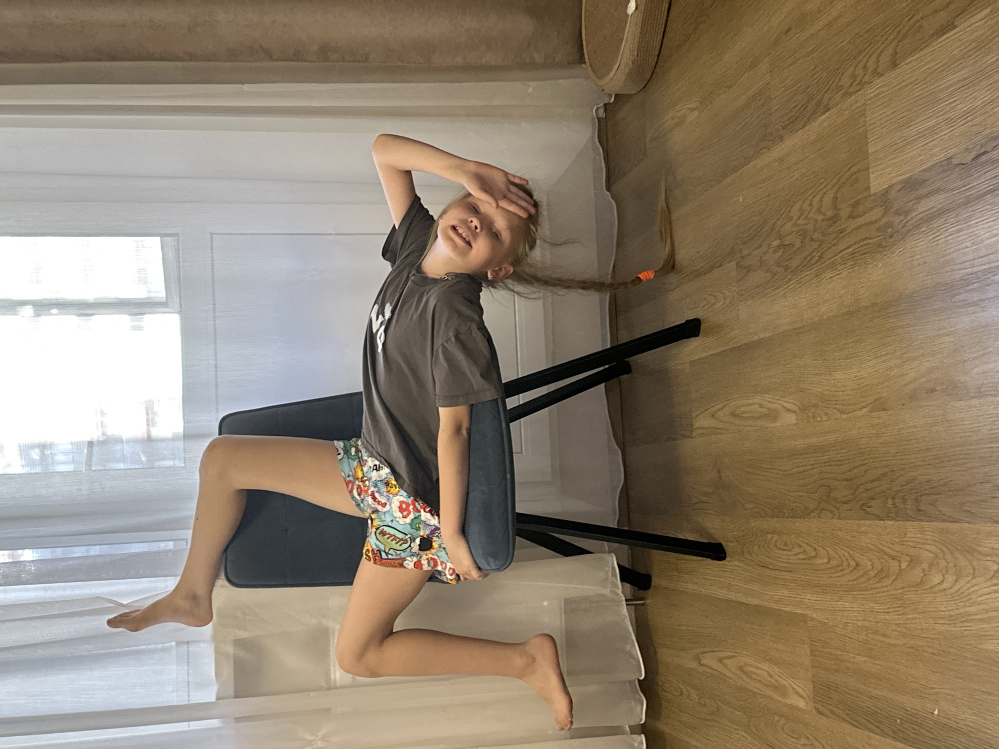 A young girl with blonde hair in a single braid, tied with an orange band, is sitting on a dark blue upholstered chair. She is wearing a dark grey t-shirt and colorful shorts with a comic book print. Her left arm is raised, with her hand on her forehead, and she is smiling. Her legs are bent, with her left foot resting on the floor and her right leg extended outwards. The background shows a window with white curtains and wooden flooring. The scene appears to be indoors, possibly a home. The lighting suggests daytime. The girl's expression conveys a sense of playfulness or amusement.