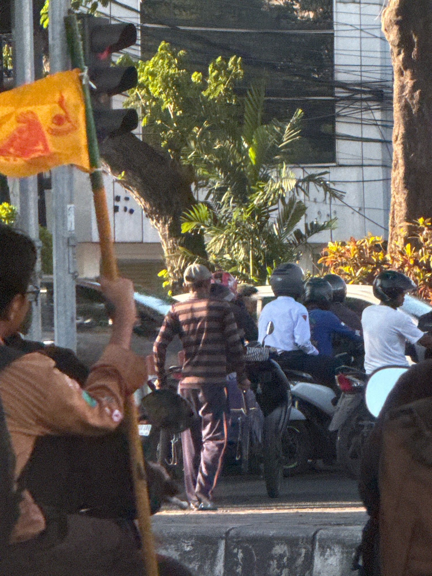 In Surabaya, Indonesia, on a street corner, a yellow flag with a red emblem is attached to a traffic light pole. Three men on motorcycles are visible. The man in the foreground is wearing a striped shirt and a grey helmet. The second man, in the middle, is wearing a white shirt and a black helmet. The third man, on the right, is wearing a white t-shirt and a black helmet. A building with a glass facade is in the background, and lush green trees are on the left side of the image. The scene suggests a busy urban environment.